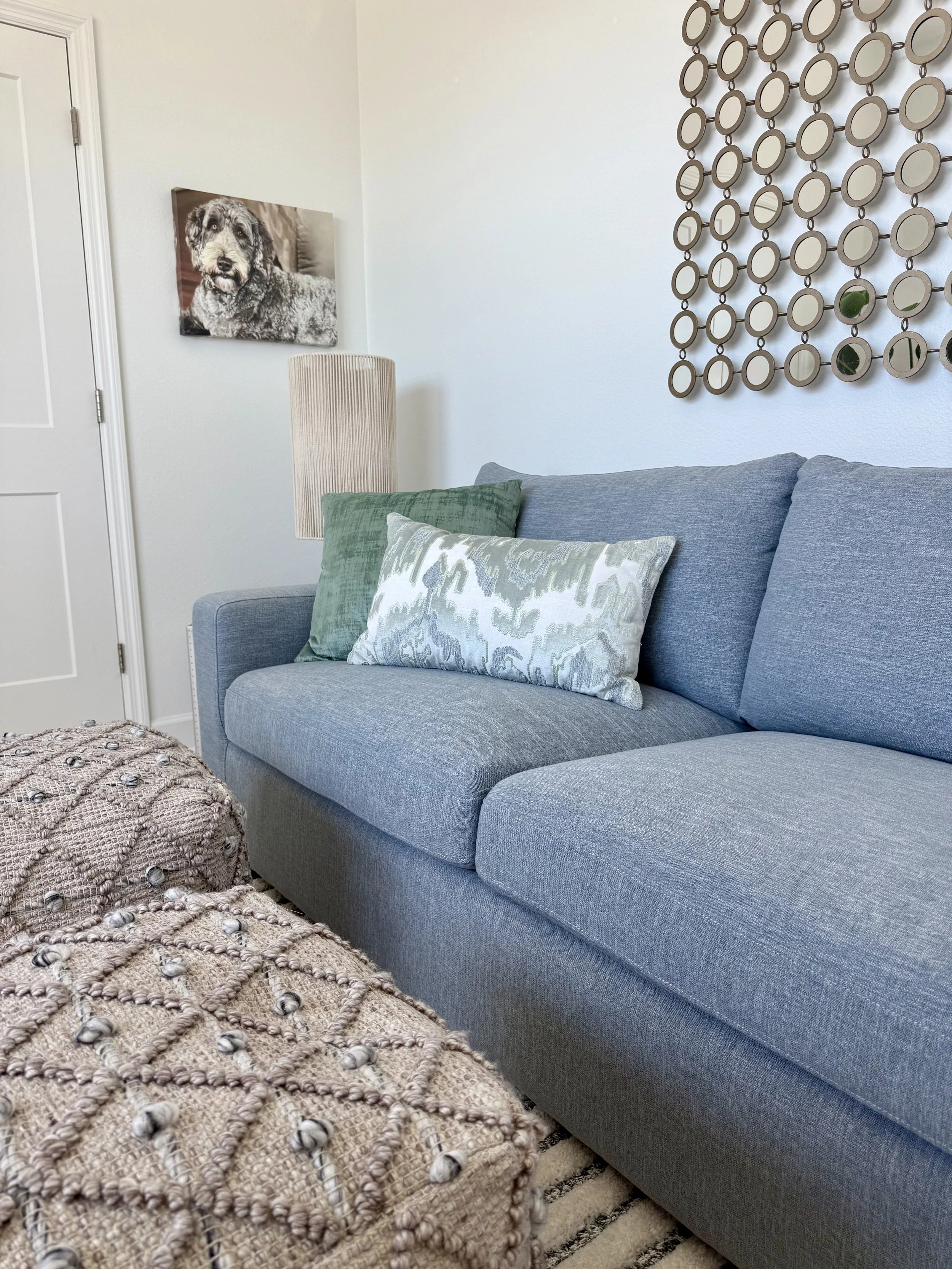 Mid-century coastal living room detail in Harbor Haven featuring a soft blue sofa, layered textured pillows, woven ottoman, sculptural wall décor, and relaxed beach-inspired styling.
