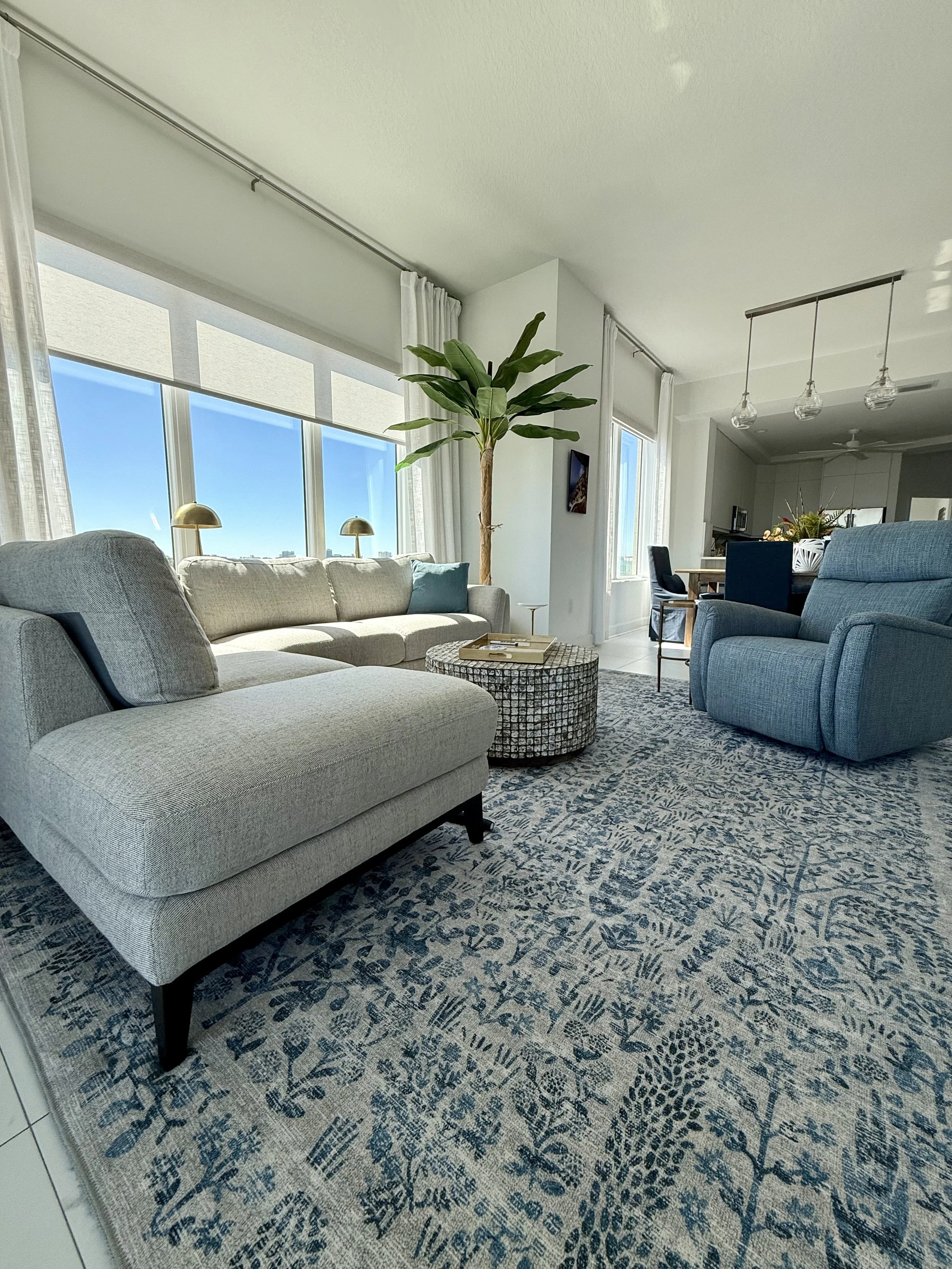 Open concept coastal living space with soft neutral furnishings, patterned blue rug, natural textures, and abundant natural light.