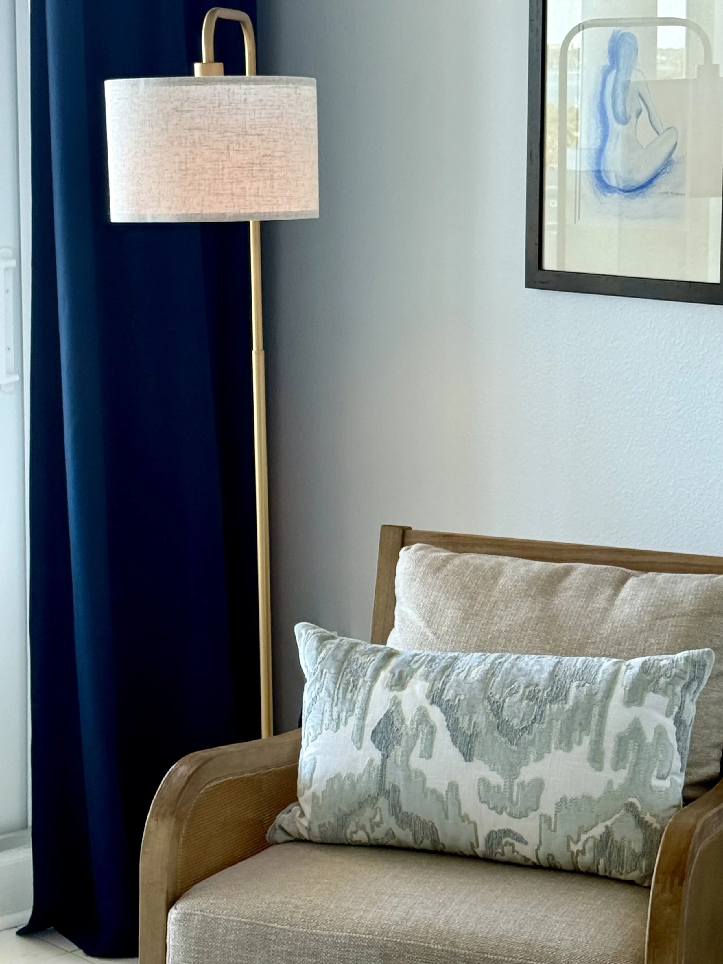 Bedroom reading corner with textured accent chair, layered pillows, brass floor lamp, and rich blue drapery.
