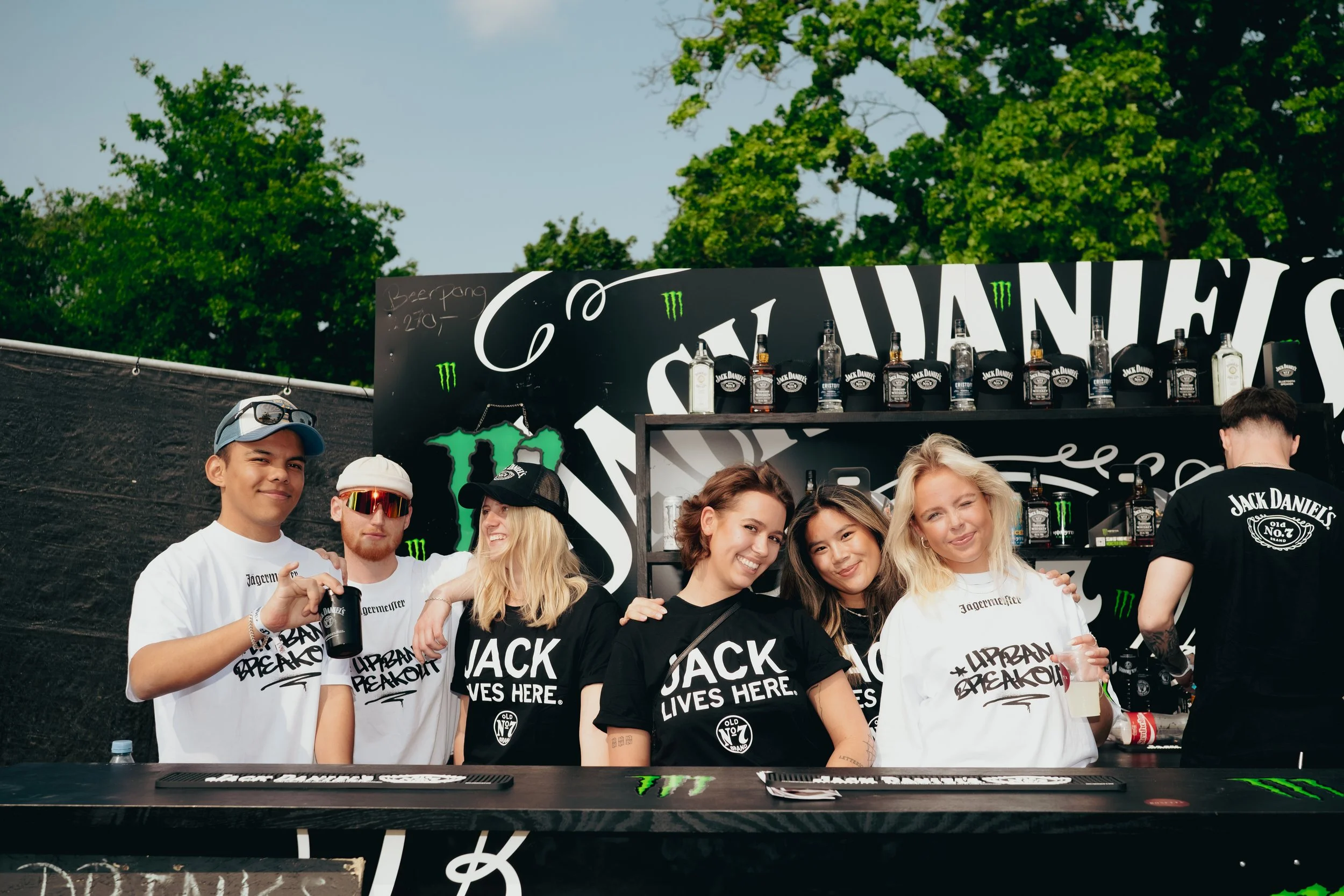 En gruppe unge mennesker står bag en bar med Jack Daniel's branding, nogle smiler og holder drikkevarer, med en sort baggrund og grønne logoer, udendørs med grønne træer i baggrunden.