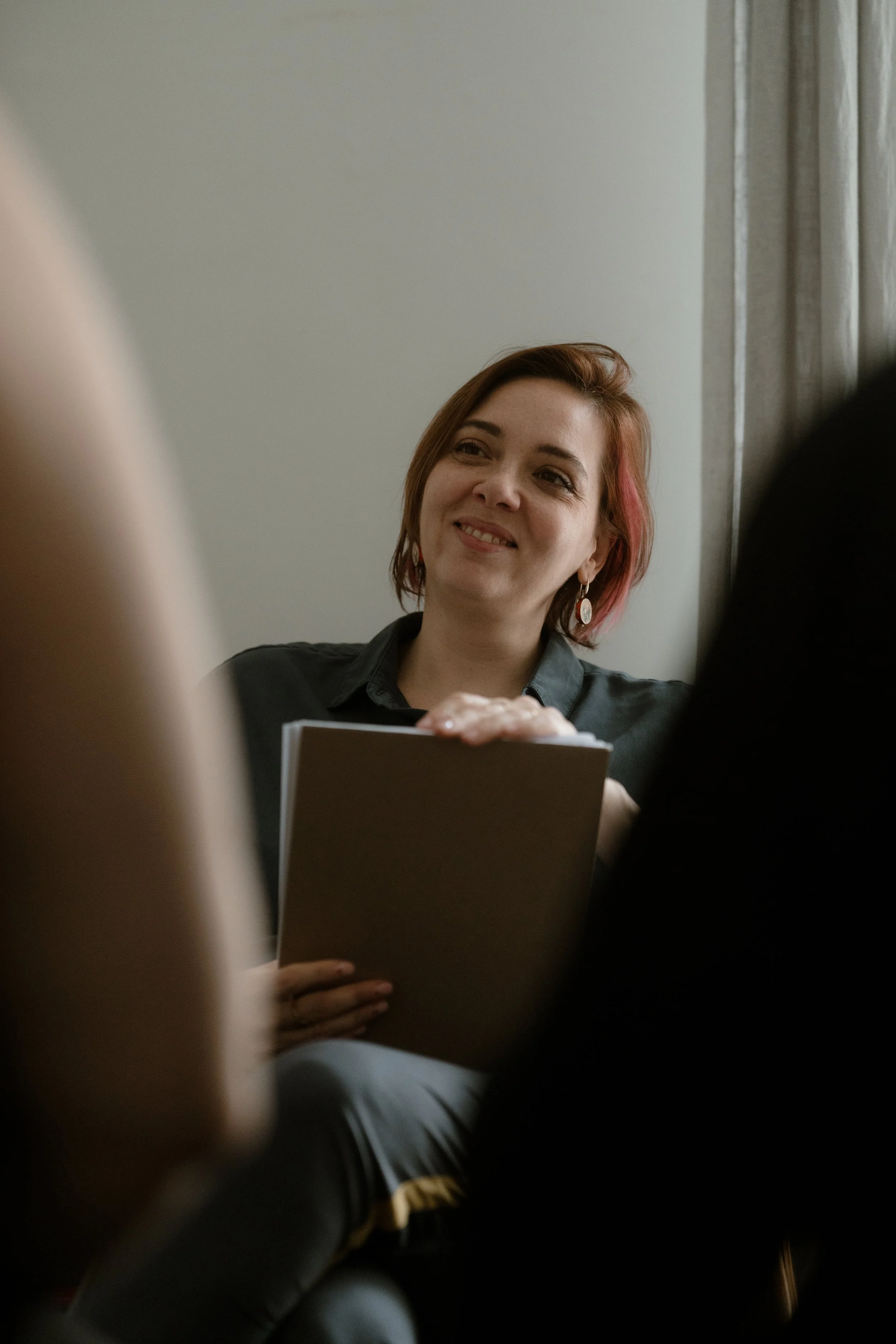 A woman with reddish-brown hair and earrings smiling, holding a book, with blurred people in the foreground and a plain wall behind her.