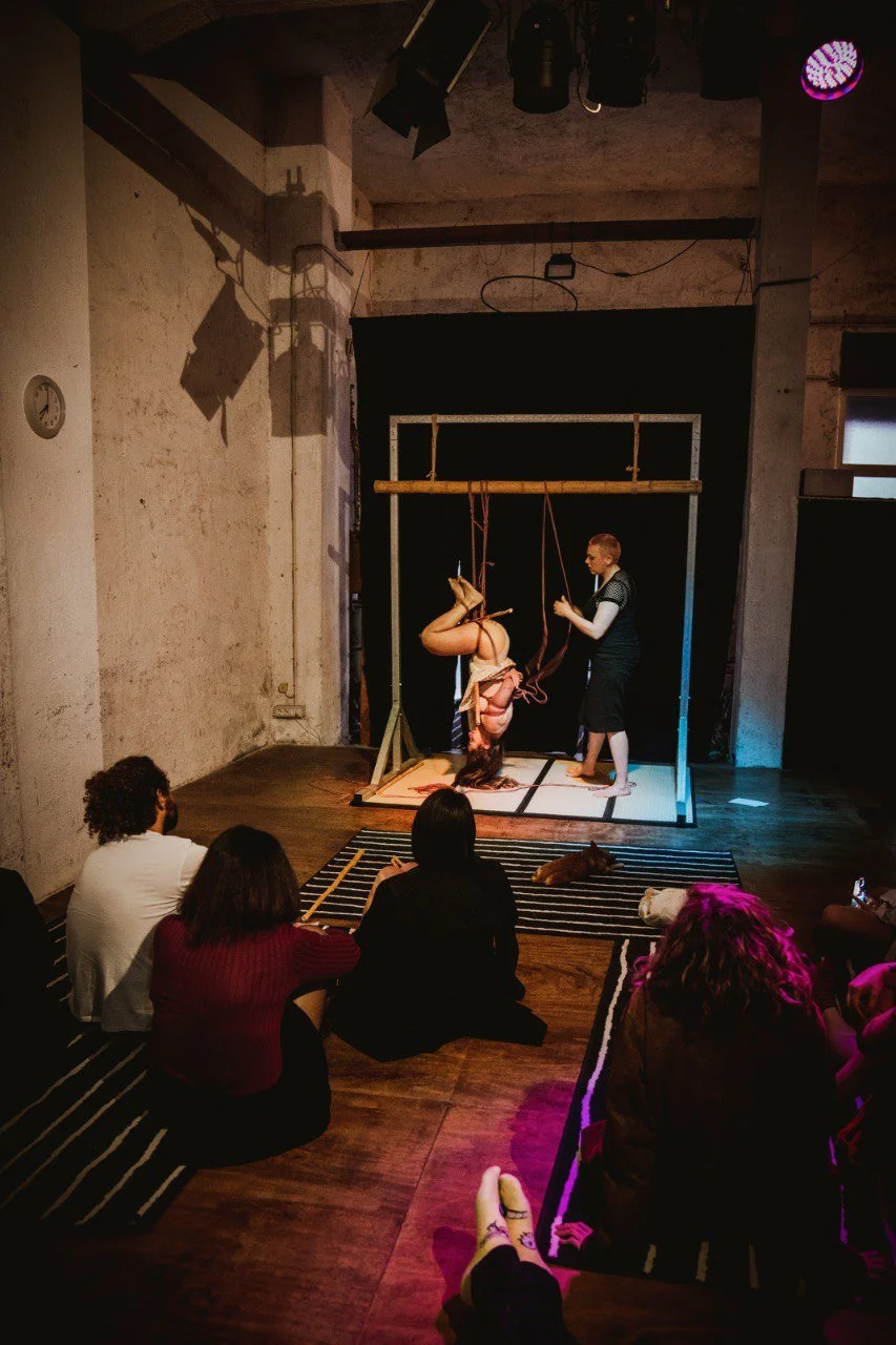 A circus or performance art scene with a woman performing aerial silks while upside down, and a man standing next to her on a stage. Audience members are sitting on the floor watching, including three women and one person with curly hair. The setting appears to be an indoor studio or performance space with stage lighting and black backdrop.