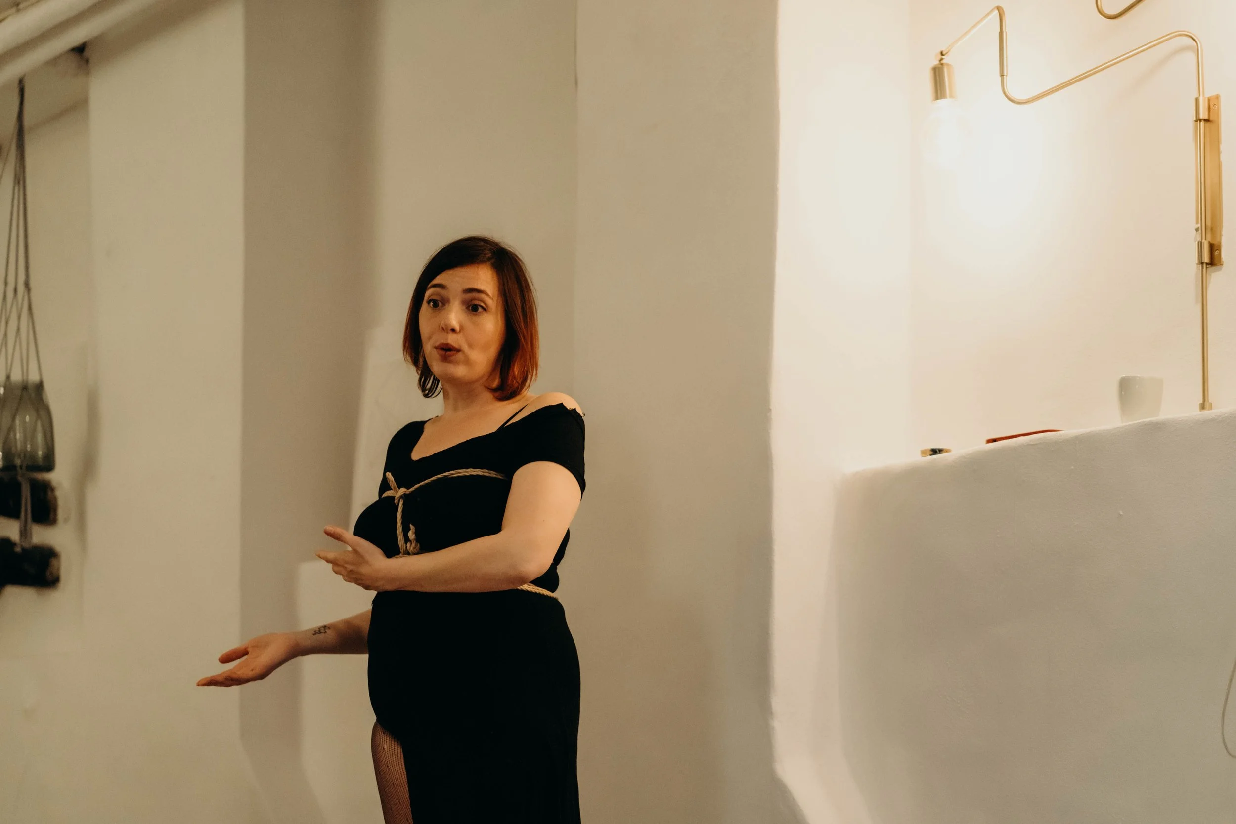 A woman with shoulder-length brown hair wearing a black dress, tied with a rope, standing indoors against white walls and a wall-mounted light fixture, gesturing with her hands.