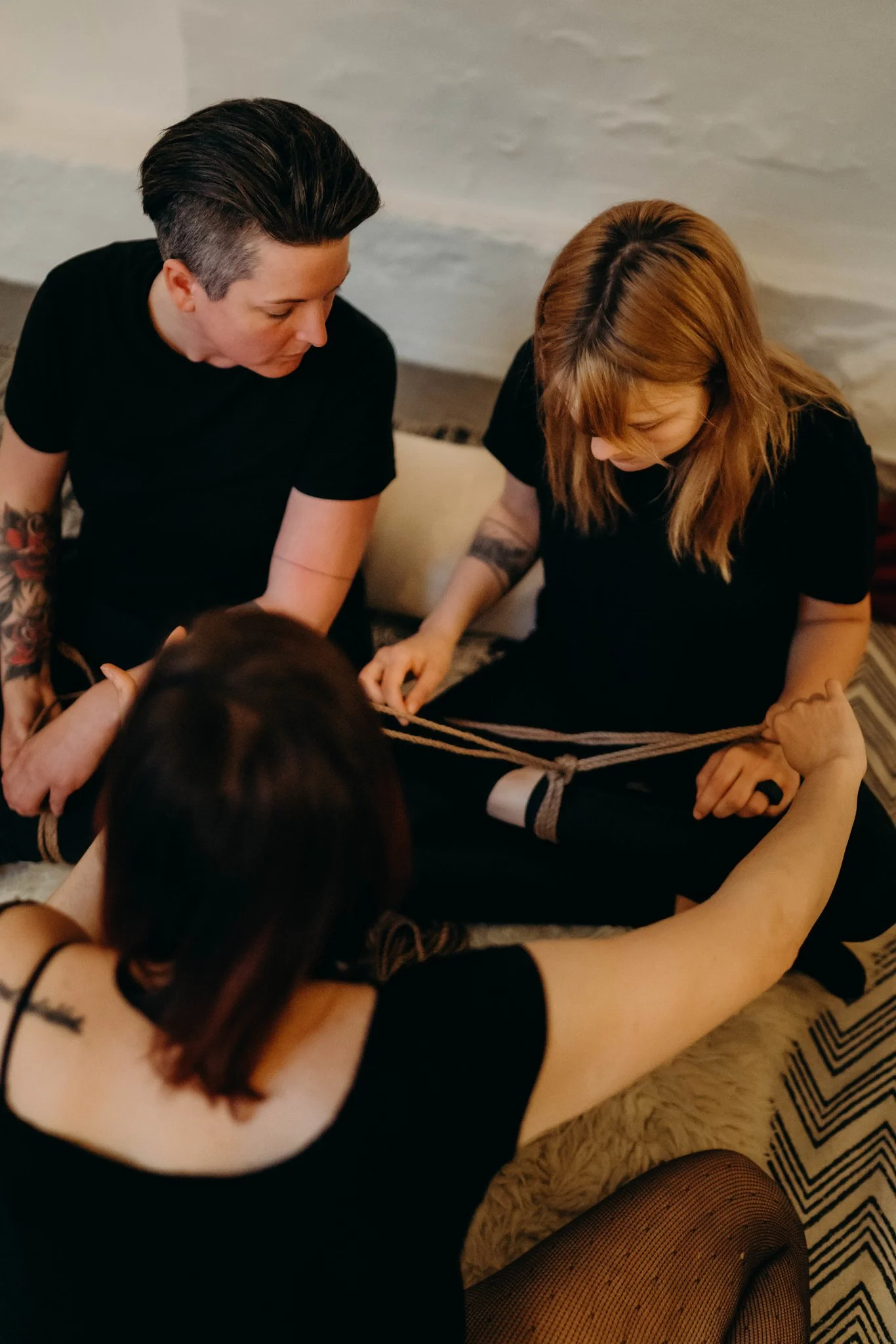 Three women, two with tattoos, are sitting on a carpeted floor. One woman has a tattoo on her right arm and black hair with a shaved side. The other woman has red hair and is holding a rope that is tied around the third woman’s ankle, who is wearing 
