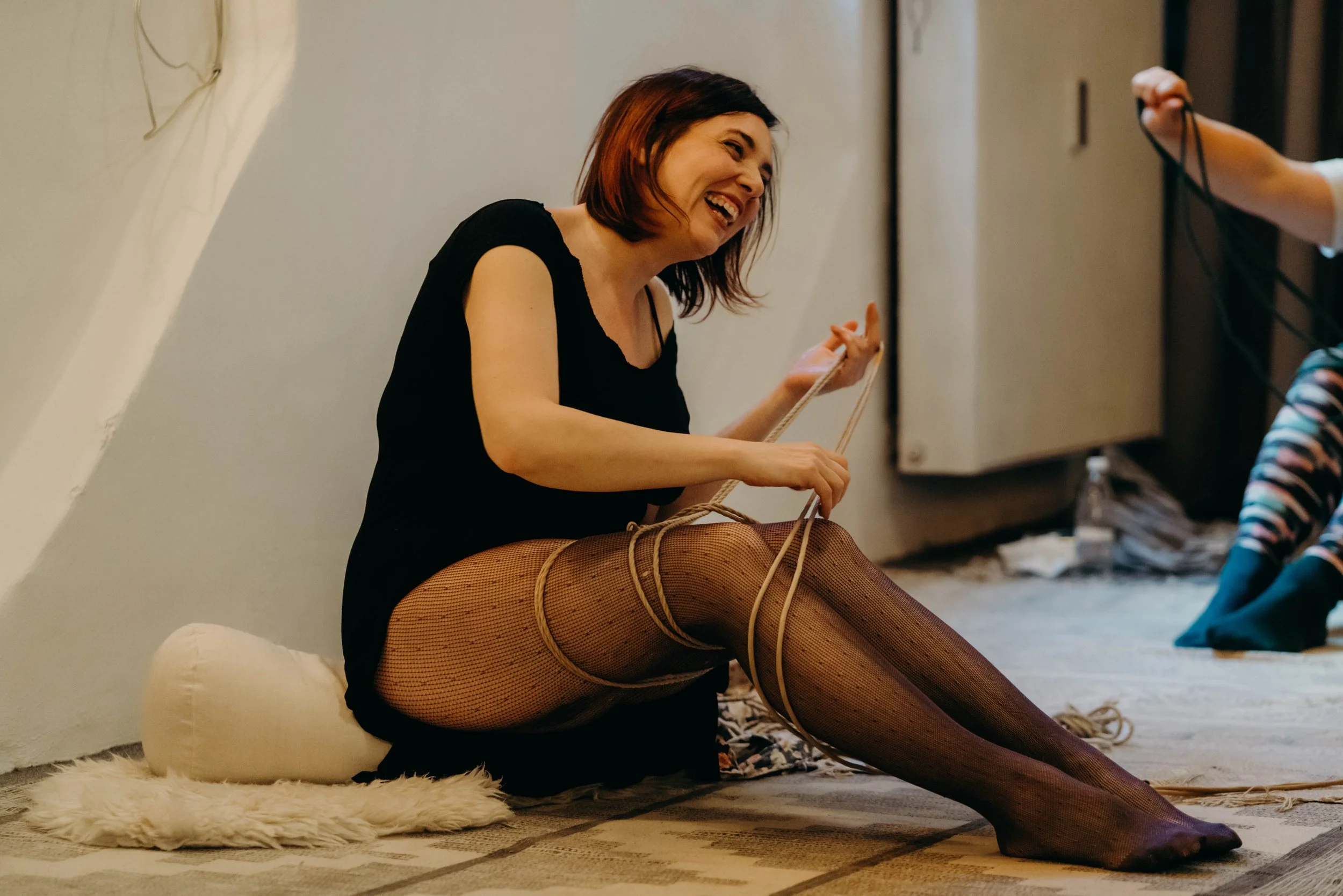 A woman sitting on the floor, laughing, with her legs tied with ropes and wearing fishnet stockings. She is holding the ropes and appears to be in a playful, joyful moment.