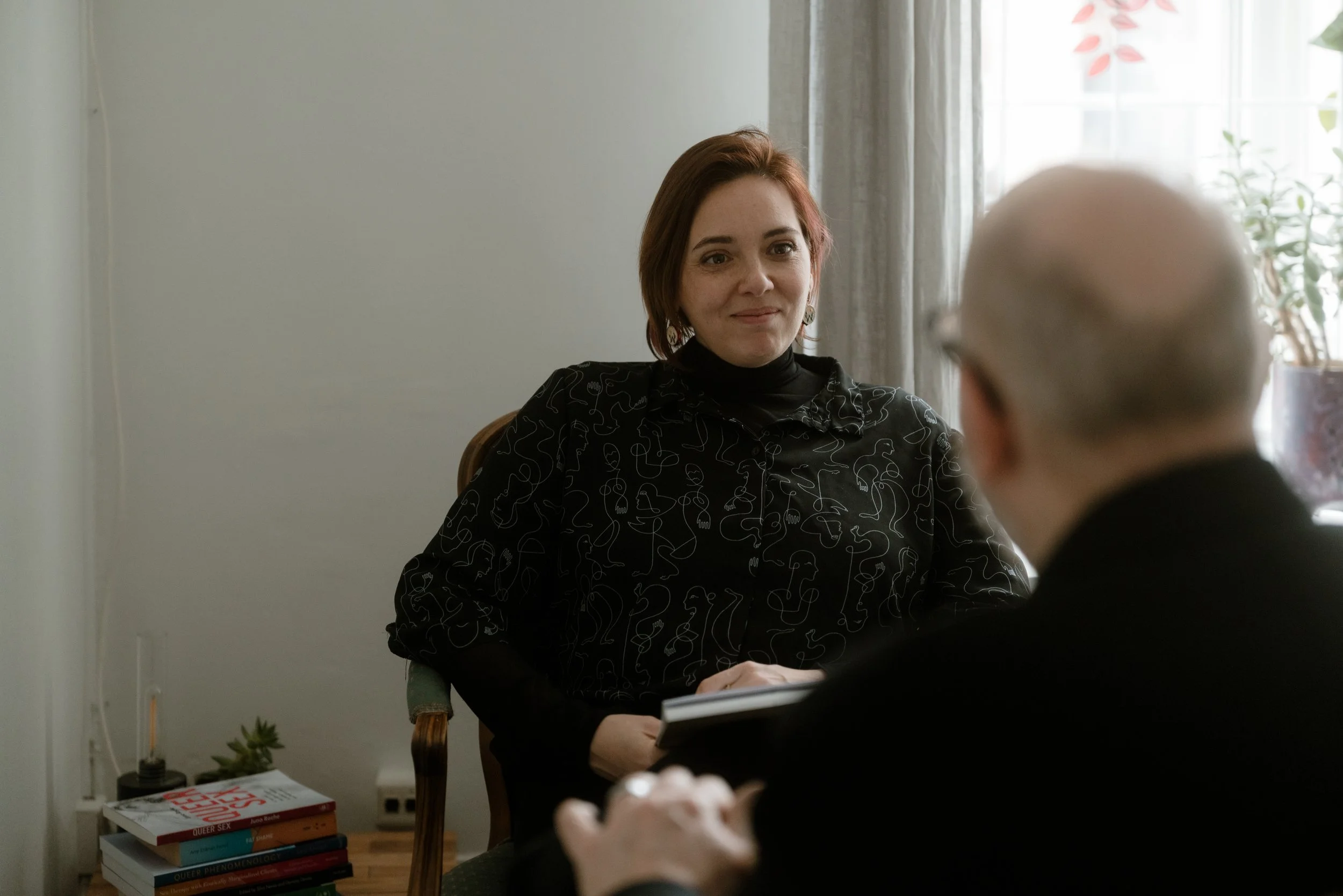 A woman with short reddish-brown hair and earrings sitting at a table during an interview, smiling at a person whose back is to the camera, in a room with a window, curtain, some books, a small plant, and a notepad.