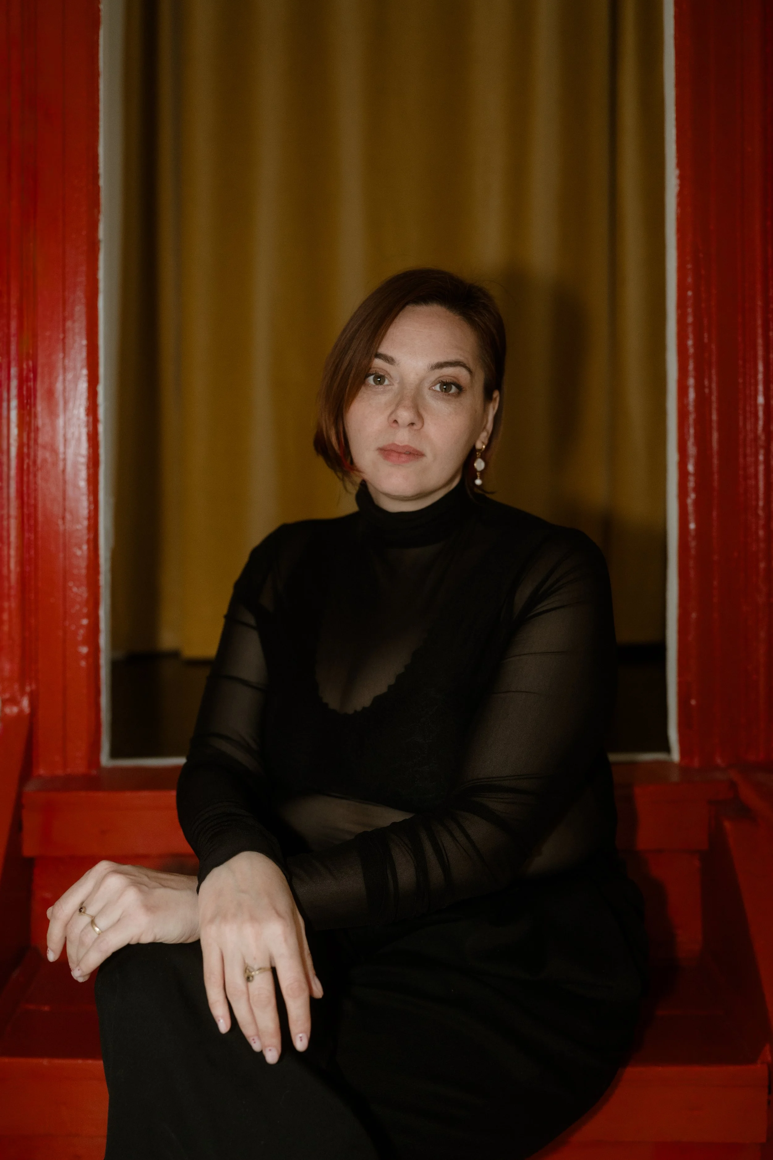 A woman with short, dark brown hair wearing a sheer black top and earrings sitting in a red wooden frame, against a background of yellowish curtains.