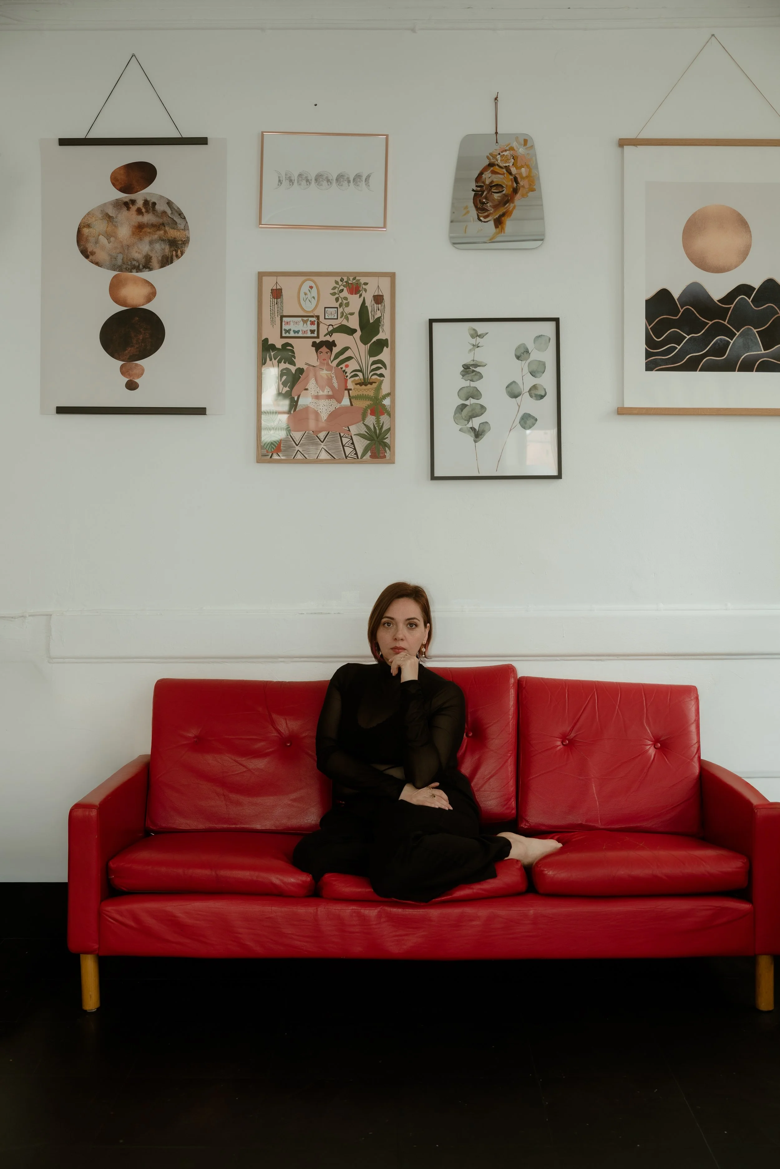 A woman with short brown hair, wearing a black long-sleeve dress, sitting on a red leather couch against a white wall with framed art and a mirror