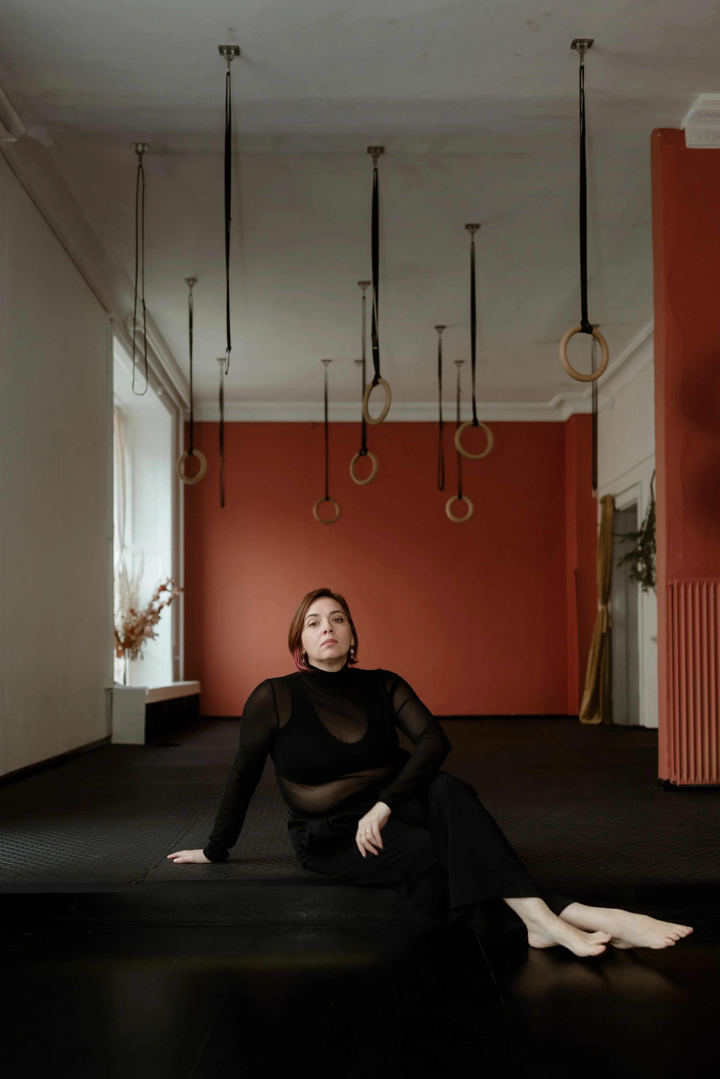 A woman dressed in black sitting on a mat in a room with gymnastic rings hanging from the ceiling and a red wall in the background.