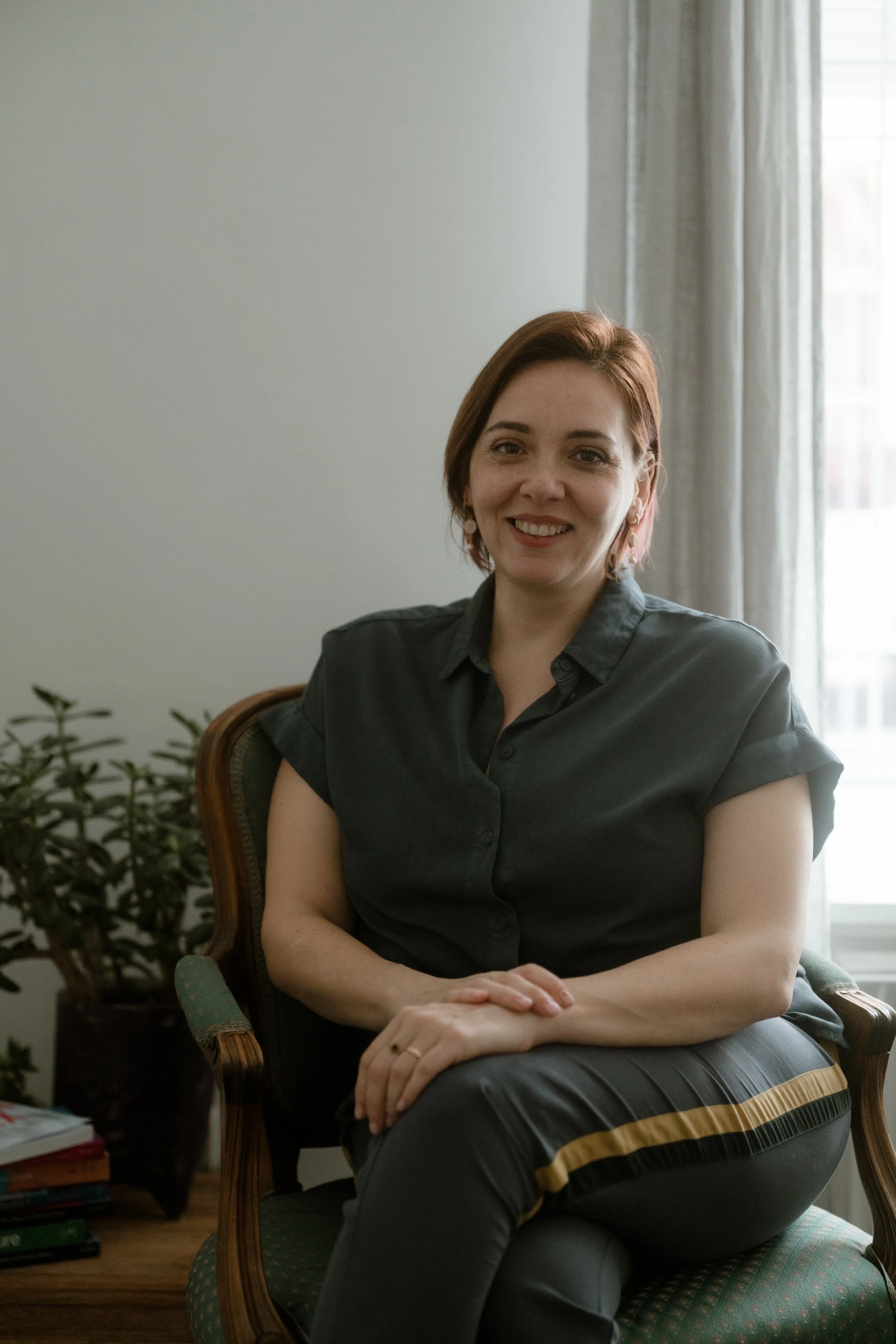 A woman with short red hair sitting on an armchair in a room with natural light, smiling at the camera, with a curtain, a window, and a plant in the background.