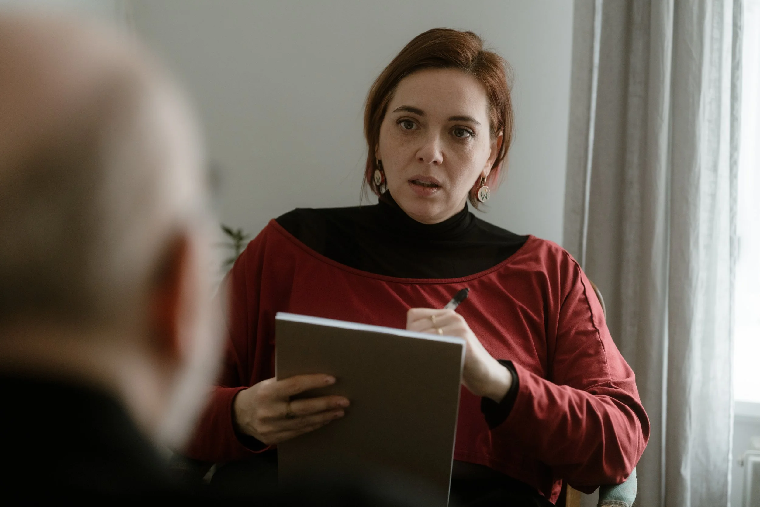 A woman with short red hair, wearing earrings and a red top over a black shirt, appears to be speaking or explaining something while holding a book or notebook, with a neutral-colored wall and curtain behind her.