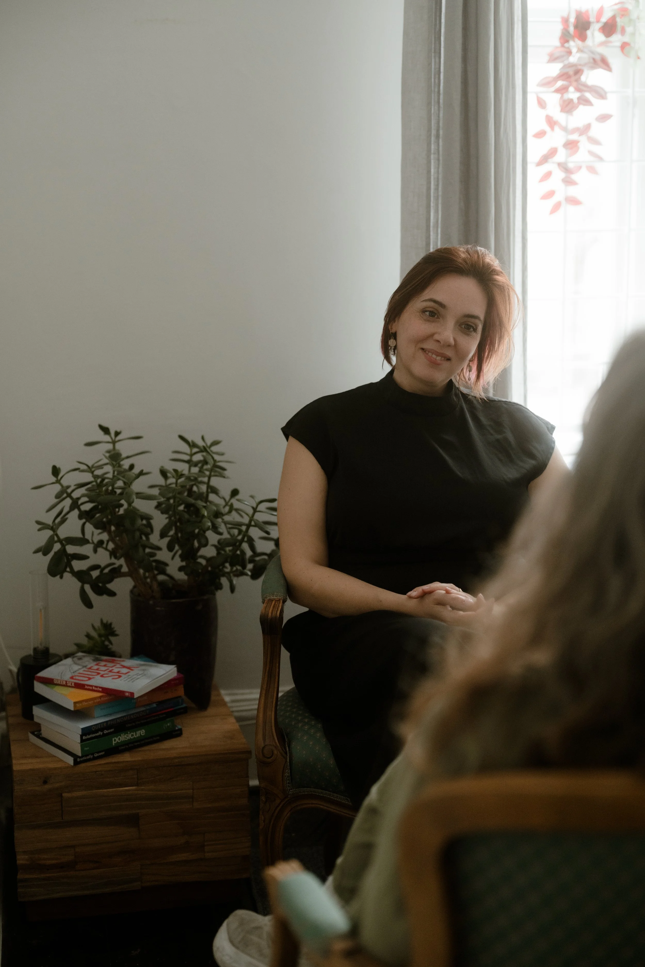 A woman with shoulder-length brown hair wearing a black off-shoulder dress and fishnet stockings, sitting cross-legged on a patterned rug, engaged in a conversation with a person whose back is turned, with visible short dark hair and a tattooed arm. They are indoors, on a decorated dark rug and lighter patterned background rugs.