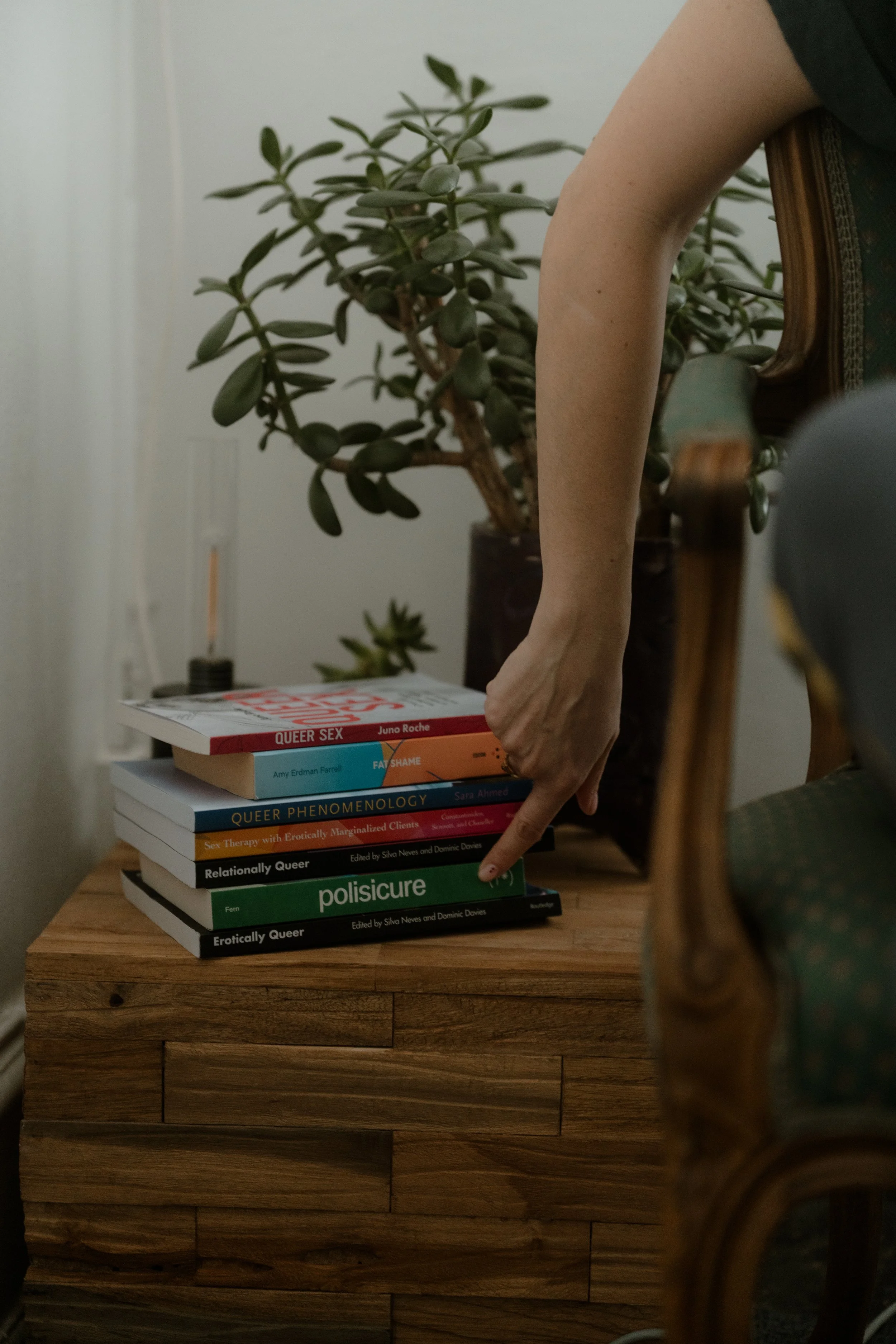 A stack of books about queer topics on a wooden table, with a hand pointing at the bottom book named "Erotically Queer." In the background, a leafy green plant is visible.