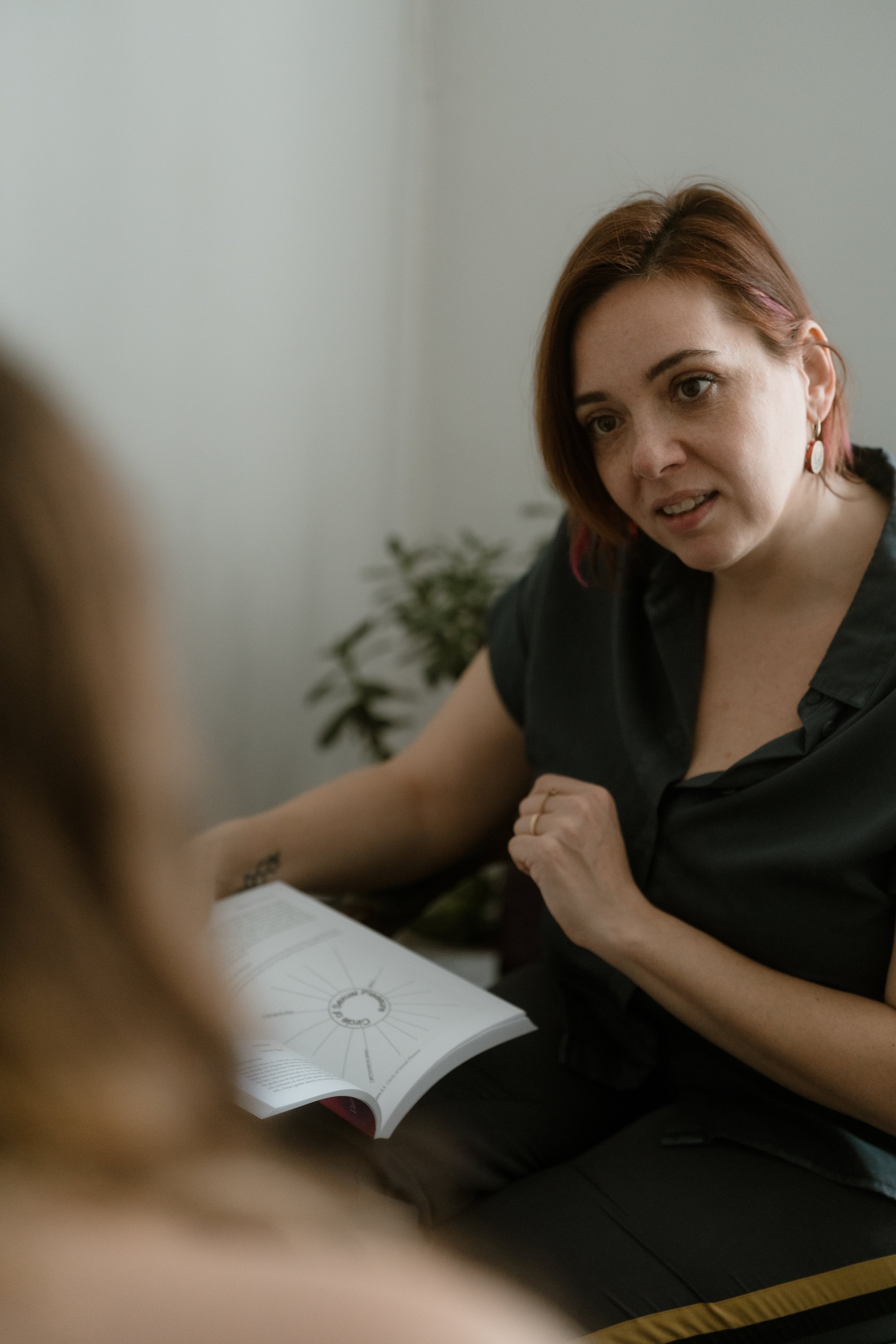 A woman with short red hair and earrings is sitting at a table, appearing to be engaged in a conversation, with an open book or magazine in front of her. There is a plant in the background.