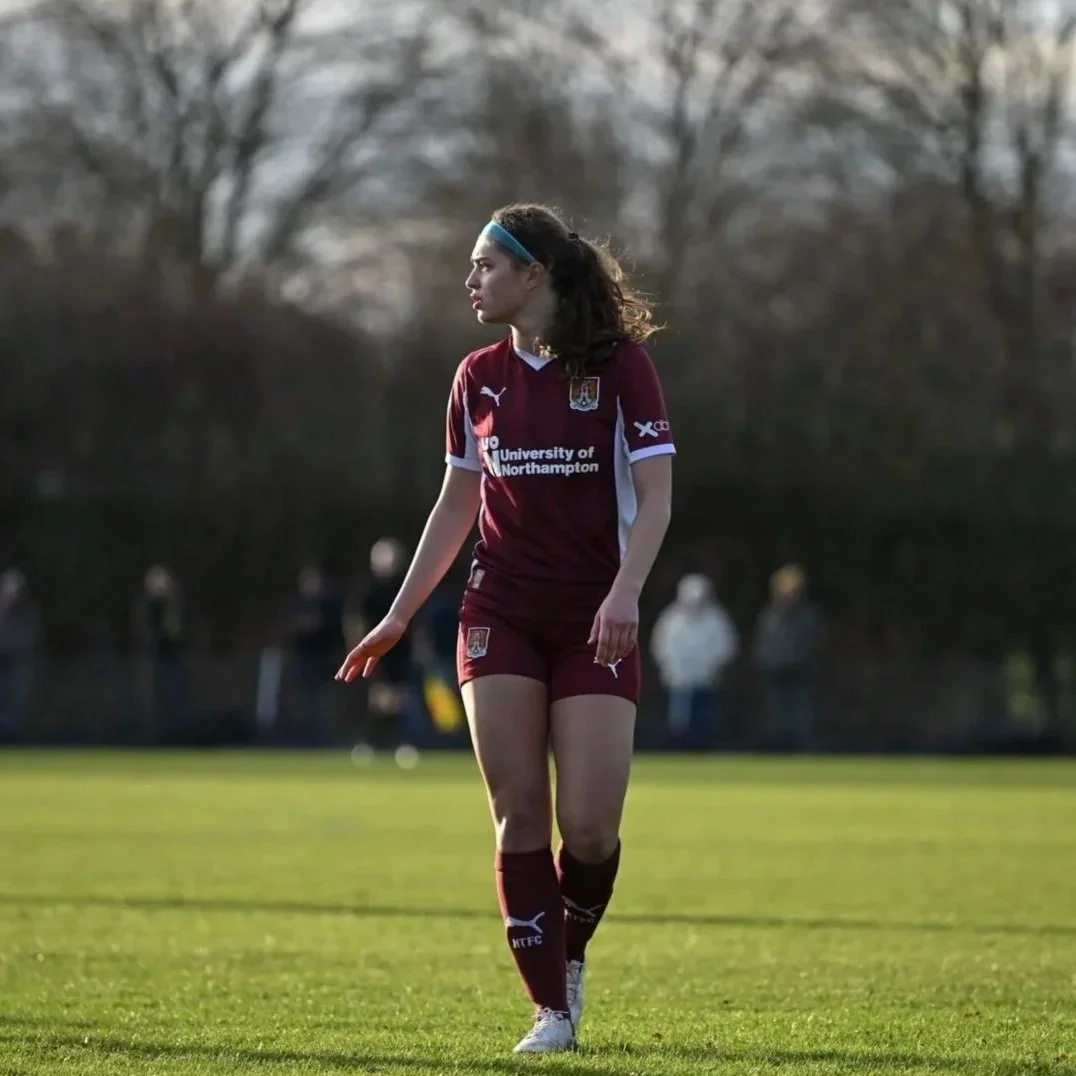 A female soccer player in a maroon uniform with 'University of Northampton' written on the front, standing on a grassy field during daytime, with trees and a few people in the background.