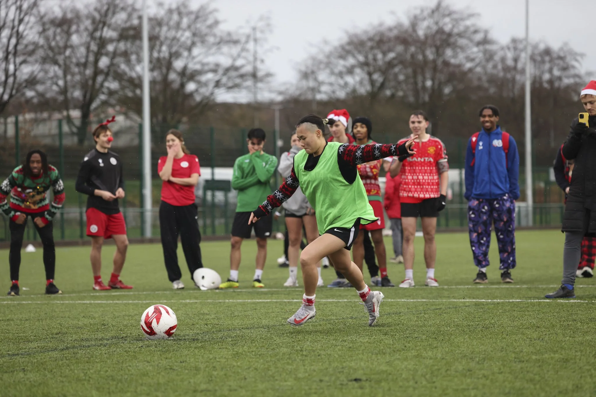 A group of people participating in a Christmas-themed soccer game on an outdoor field, with some wearing holiday sweaters and accessories.