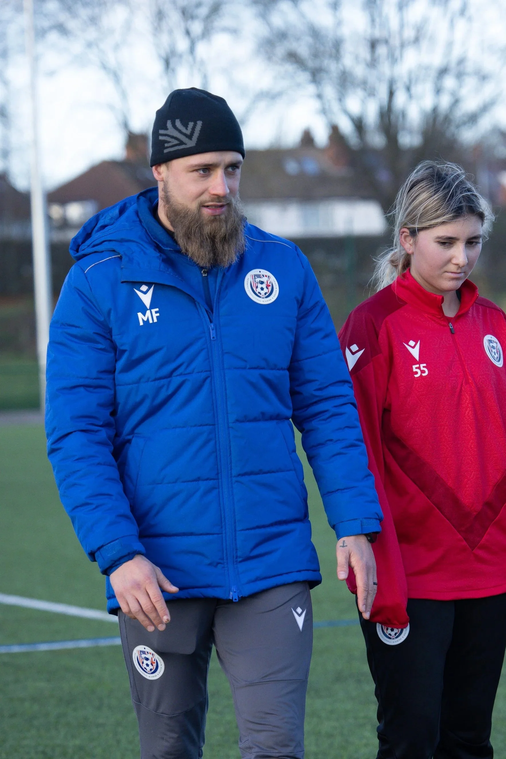 Two people standing on a soccer field, wearing sports jackets with a team logo. One is a man with a beard in a blue jacket and black beanie, the other is a woman in a red jacket.