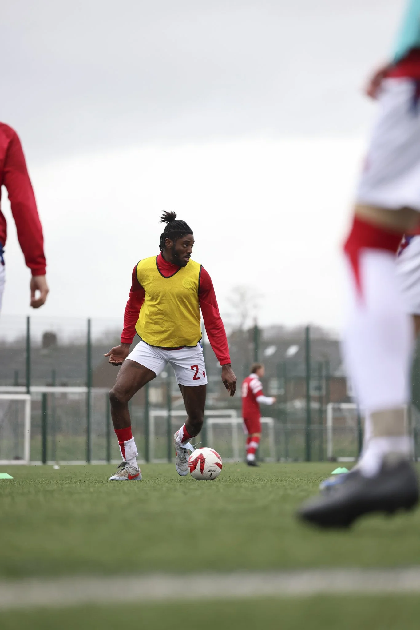 A soccer player wearing a yellow training vest over a red shirt, white shorts, and gray shoes with red and white socks, standing on a soccer field and dribbling a ball during practice.