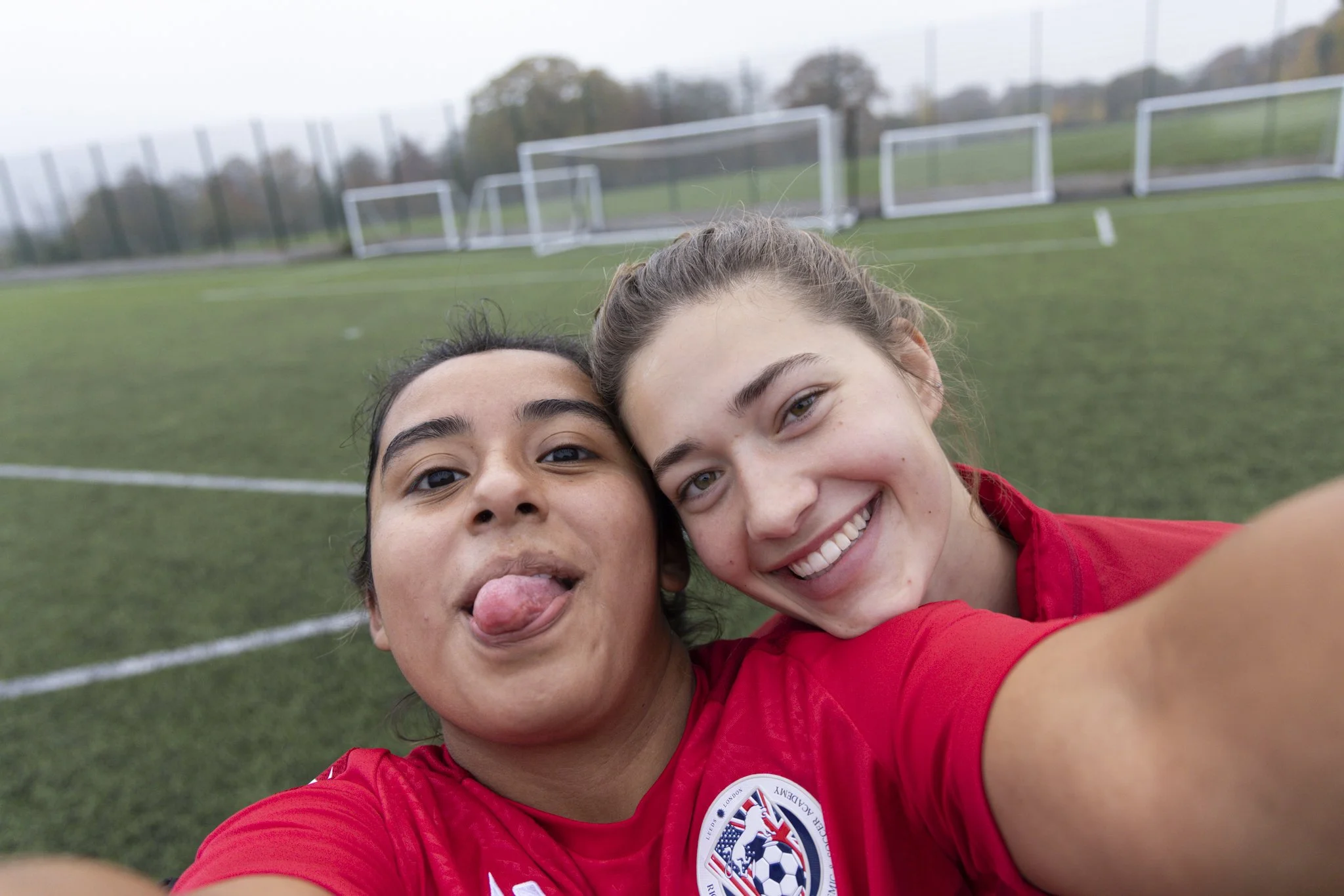 Two young women taking a selfie on a soccer field, wearing red sports jerseys. One has her tongue sticking out, and both are smiling.