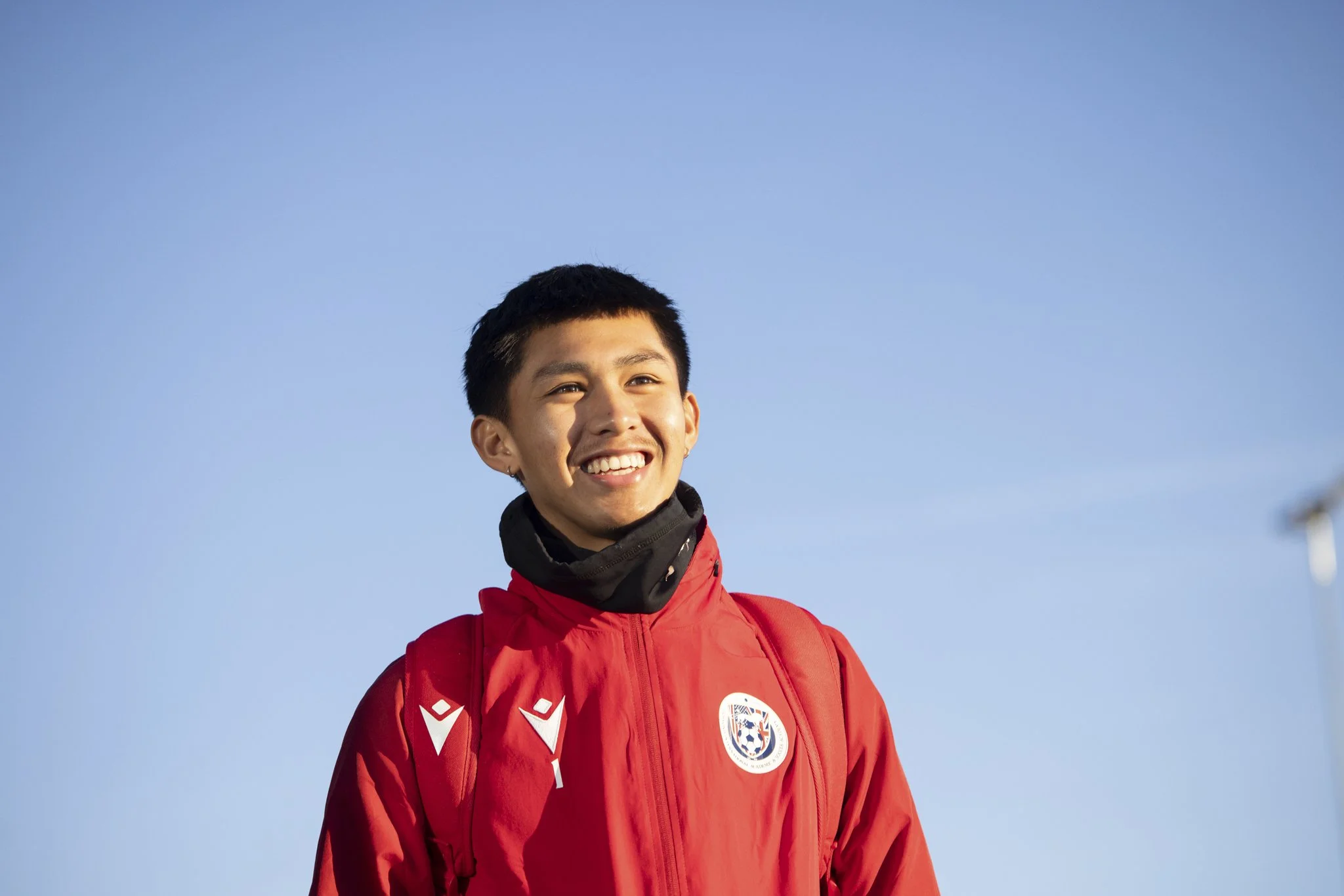 A young man smiling, wearing a red sports jacket with a badge and carrying a backpack, standing against a clear blue sky.