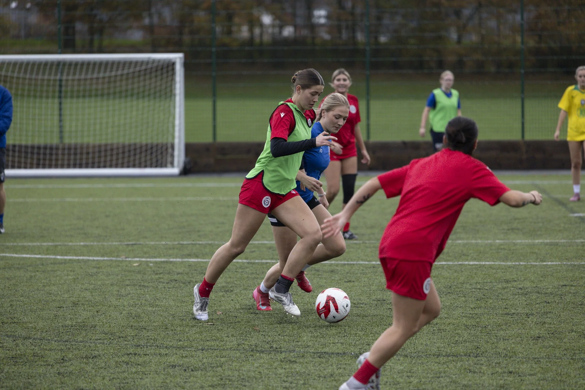 Women playing soccer on a field during daytime, with some women in red and green uniforms and others in yellow and blue uniforms, and a soccer goal in the background.