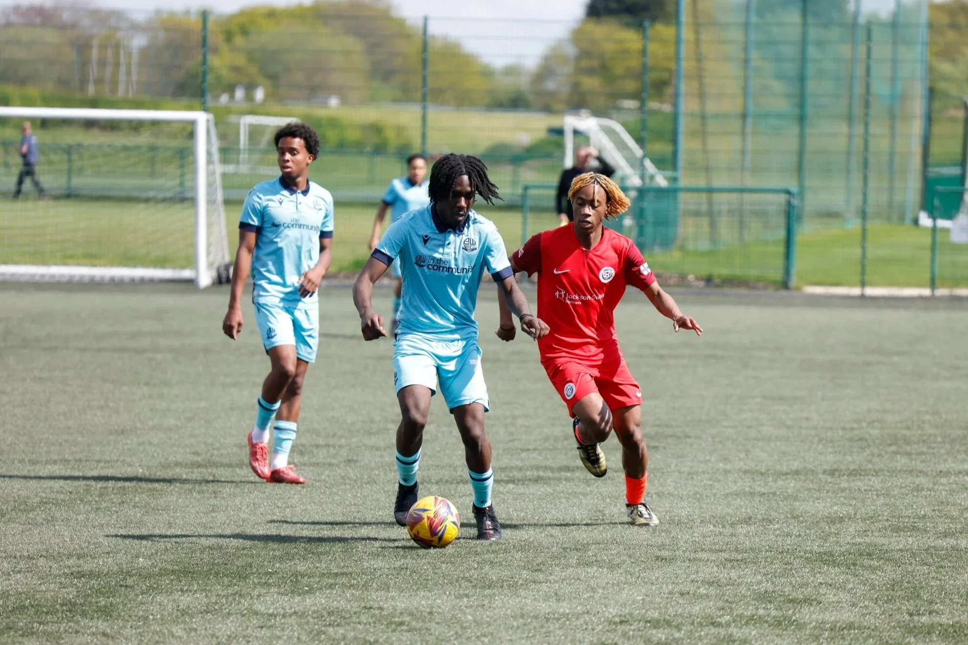 Three soccer players on a field, two in light blue uniforms and one in a red uniform, competing for control of the ball.