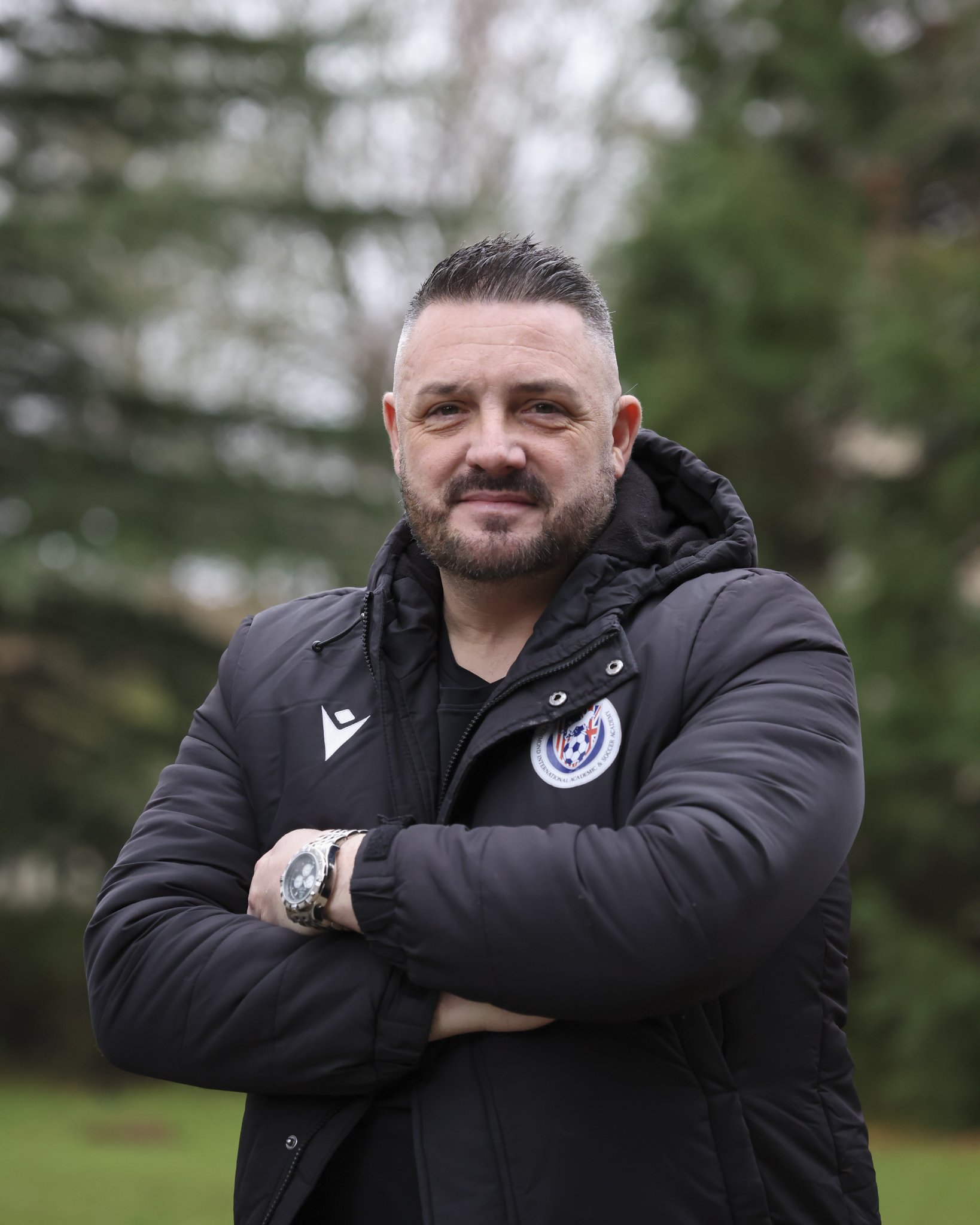 A man with short dark hair and a beard standing outdoors with arms crossed, wearing a black jacket with a sports badge and logo, and a wristwatch, with trees in the background.