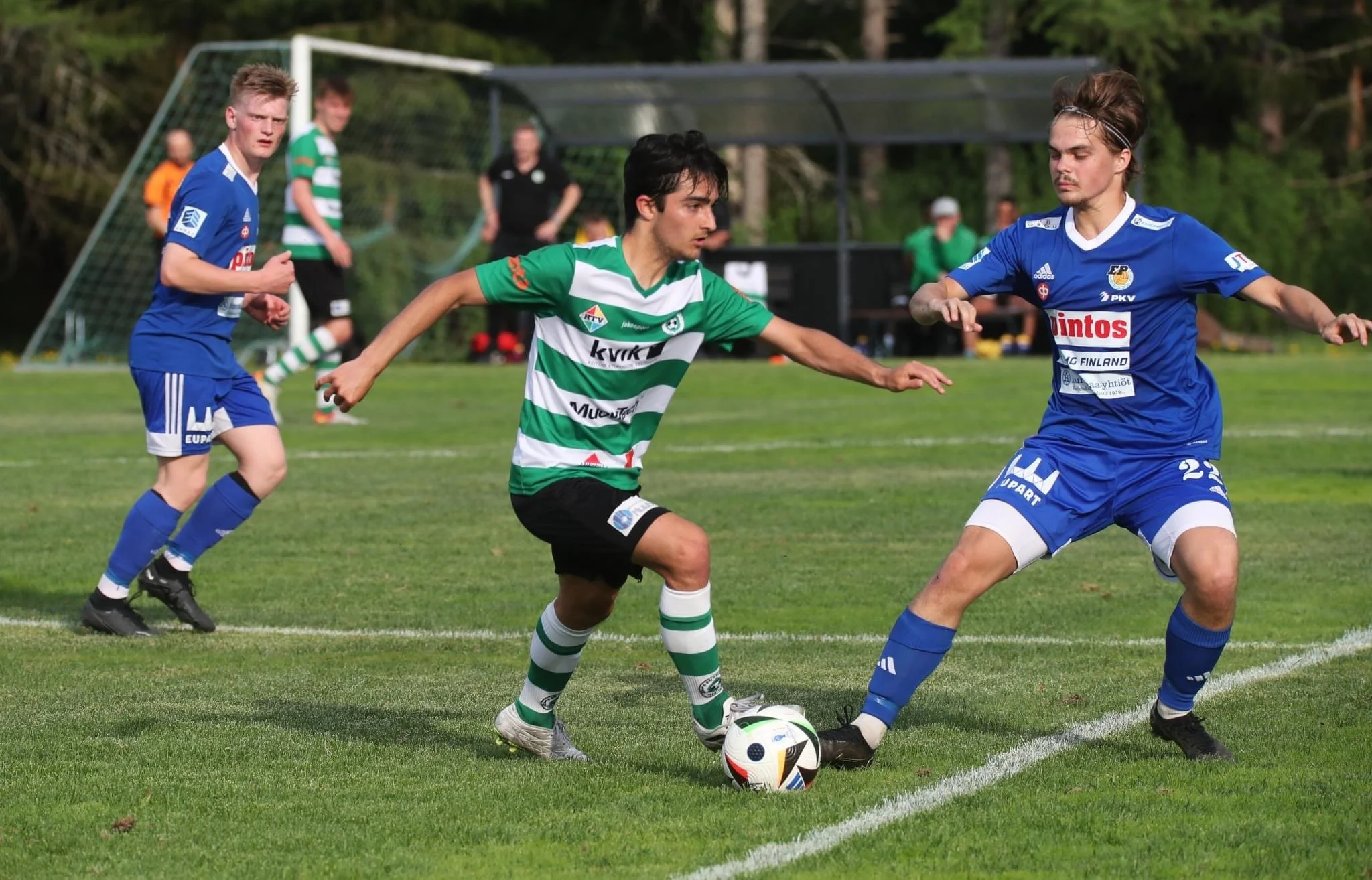 Two soccer players in blue jerseys and a player in a green and white striped jersey compete for the ball during a match on a grassy field, with other players and a goal in the background.