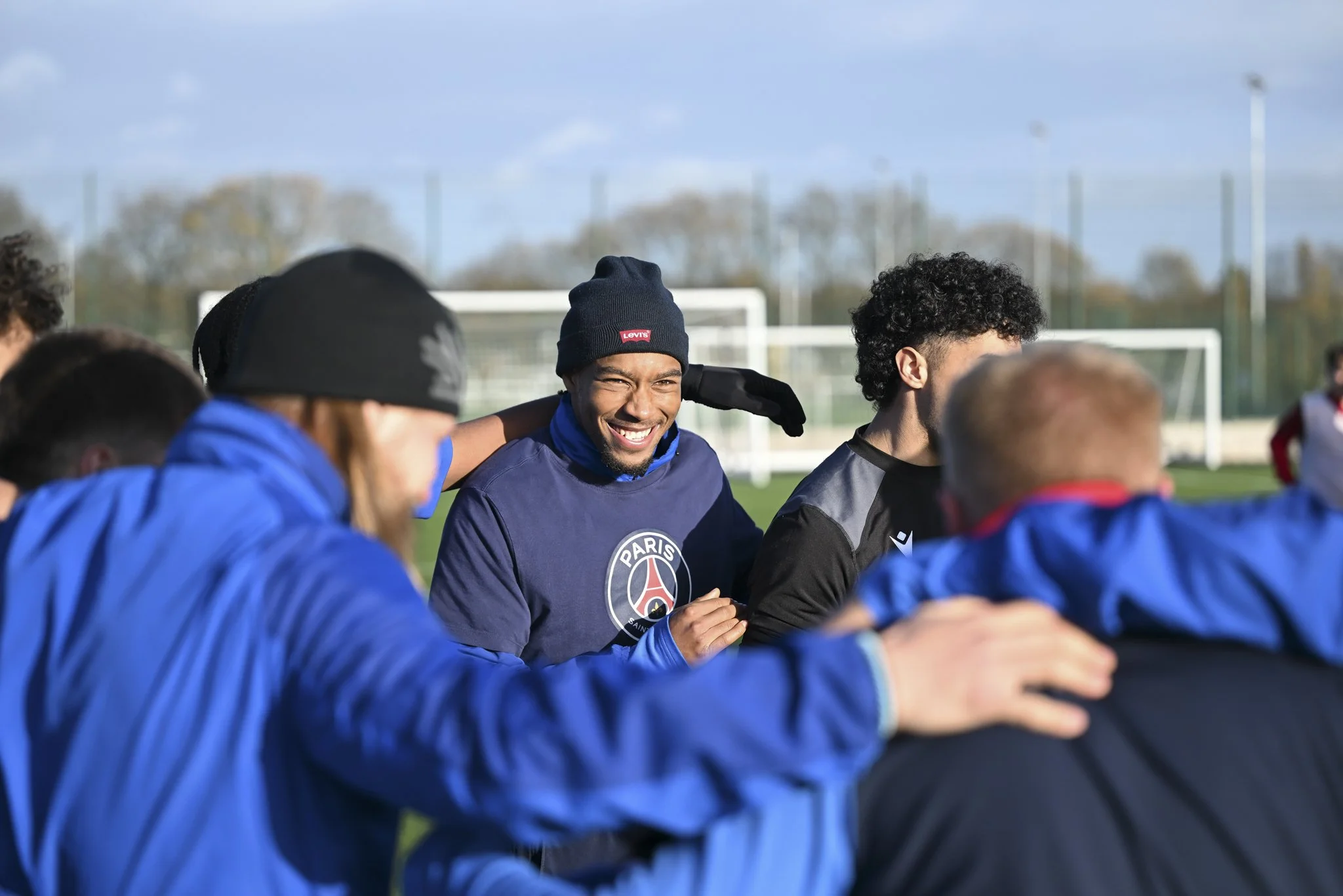 Soccer players huddled together in a team huddle on a field during daytime, smiling and wearing sportswear, with goalposts and a fence in the background.