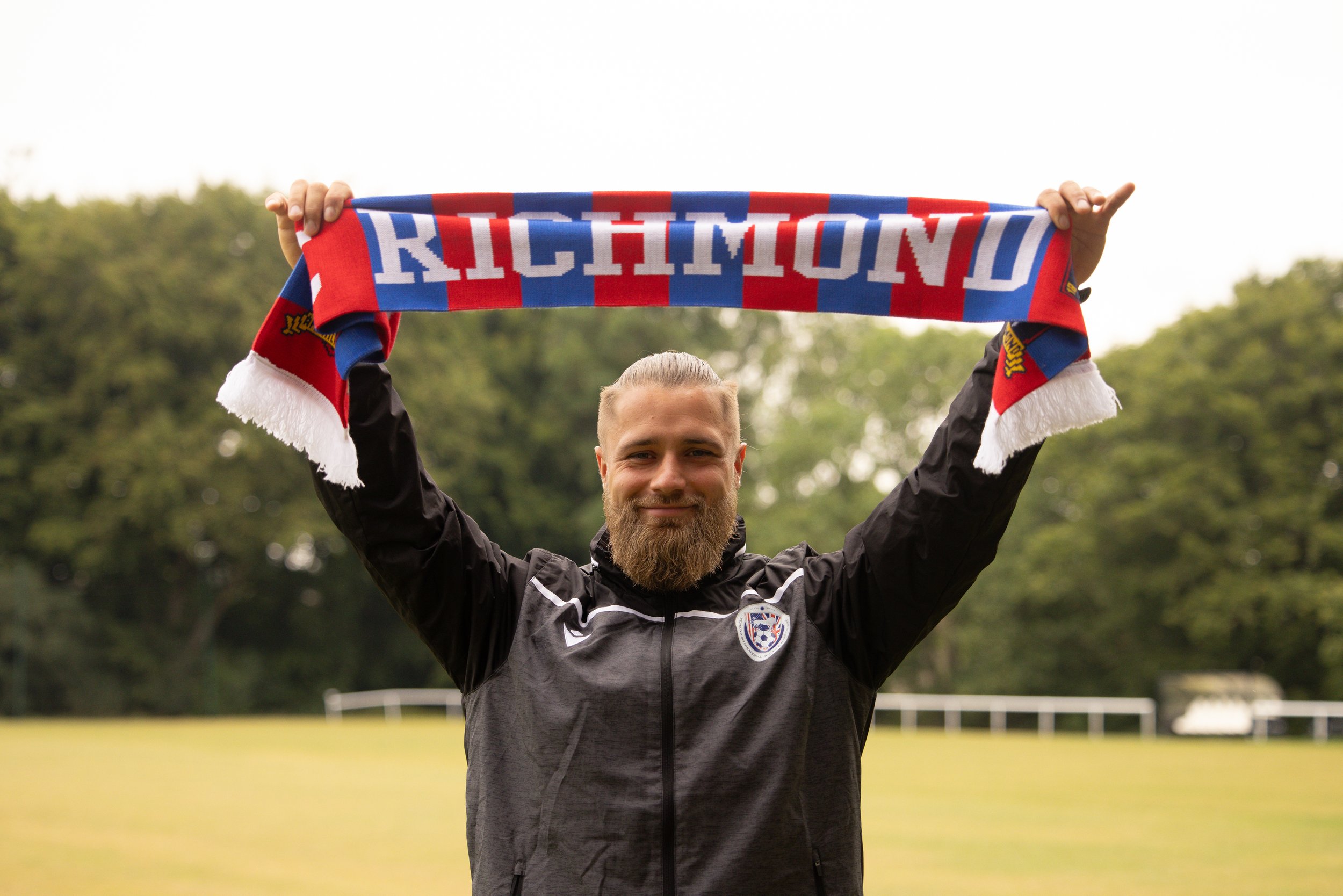 Man with a beard holding a Richmond scarf over his head outdoors on a grassy field.