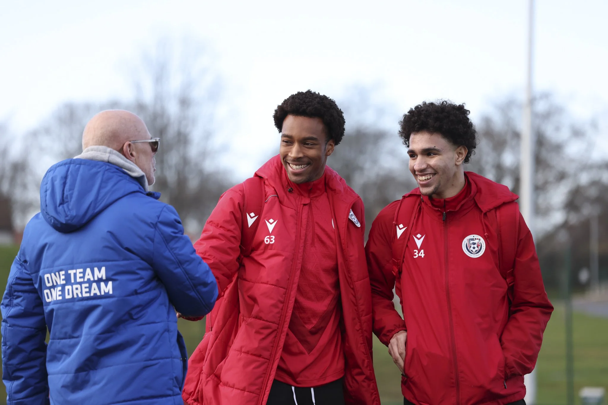 Two young male soccer players in red jackets shaking hands with an older man in a blue jacket with the slogan "ONE TEAM ONE DREAM" on the back, outdoors on a cloudy day.