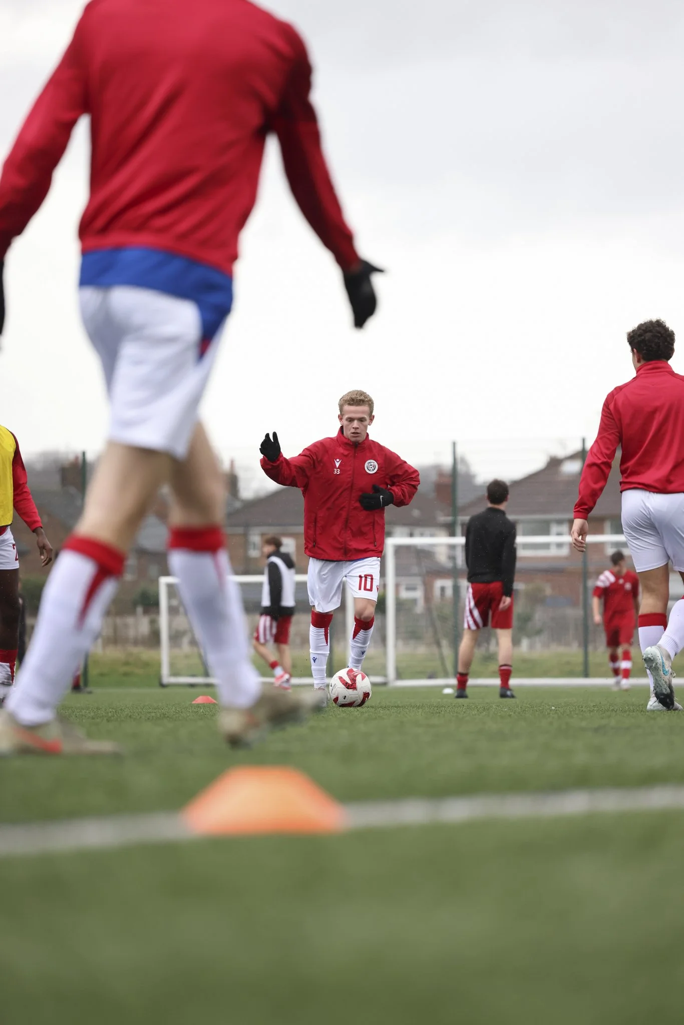 Soccer players in training on a field, with one player kicking a ball, all in red and white uniforms, overcast sky.