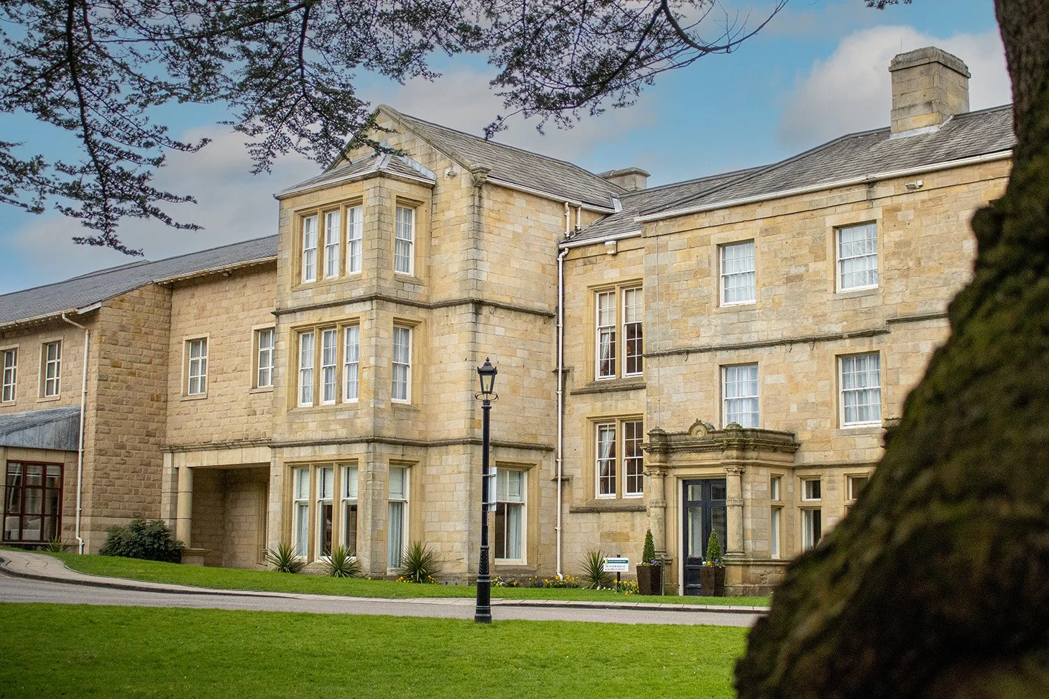 A multi-story stone building with multiple windows, a black front door, and a small porch. There's a streetlamp in front and a well-kept lawn with plants and a tree in the foreground.