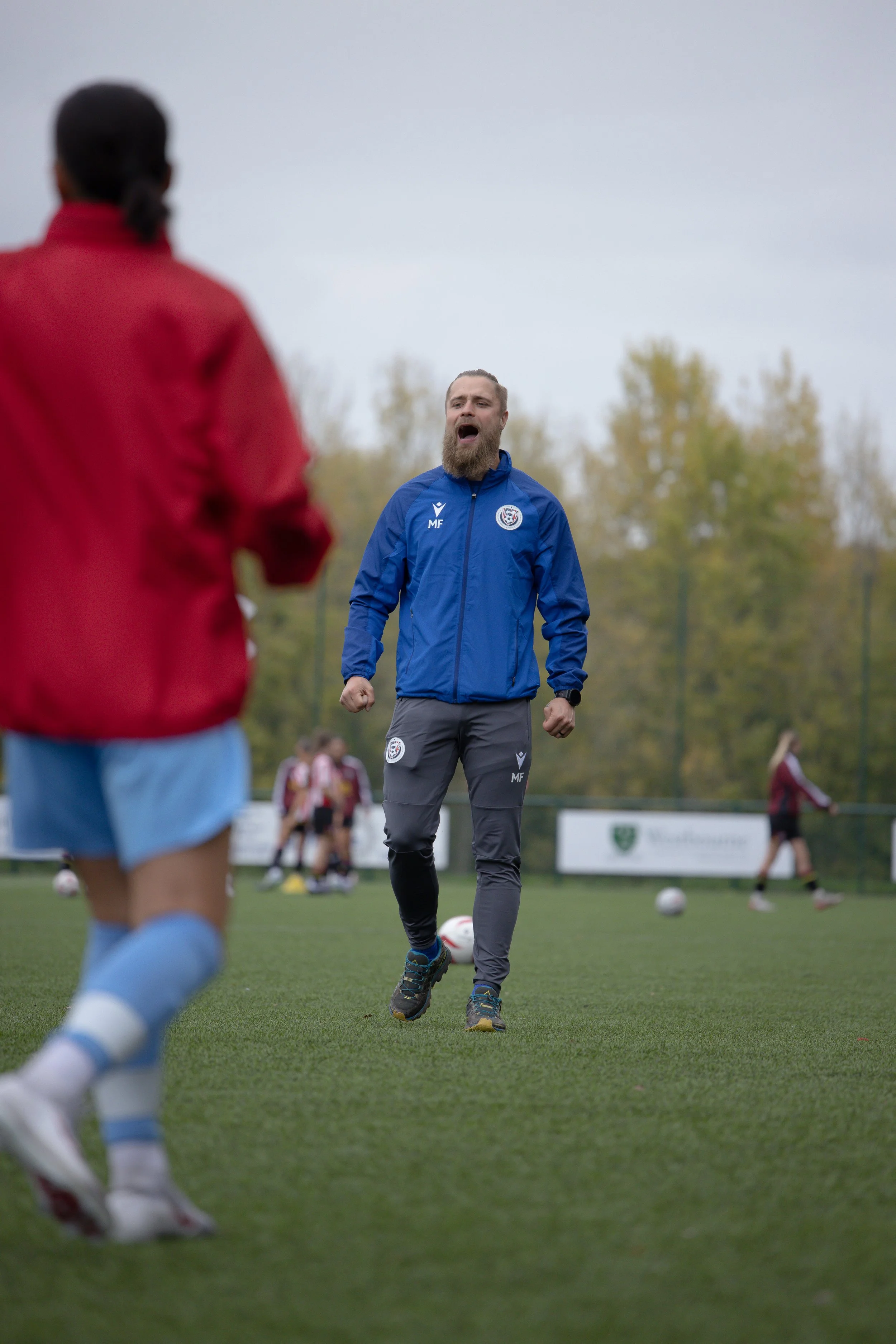 A soccer coach shouting instructions during a practice session on a soccer field, with young players in the background.
