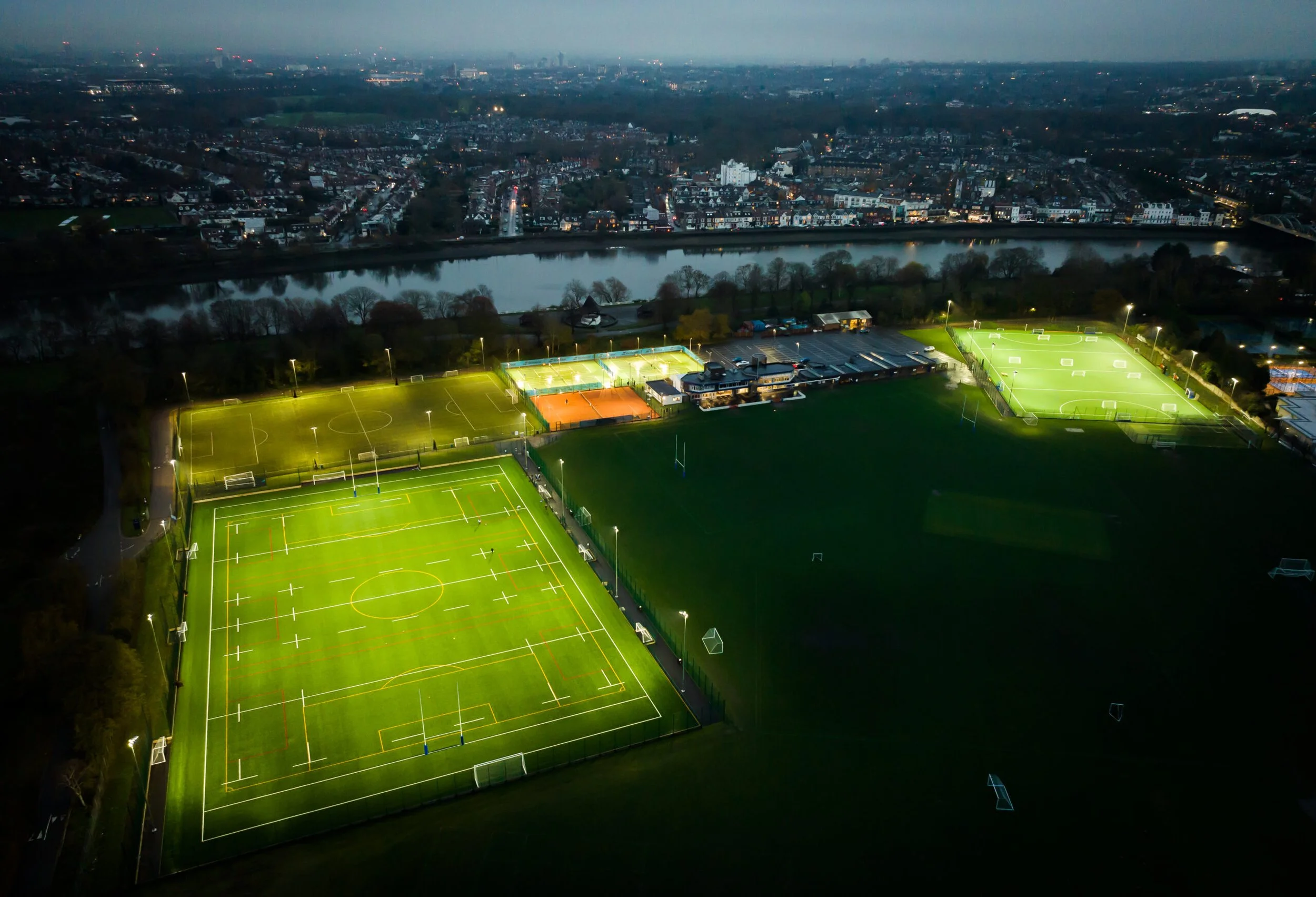 Night aerial view of a sports complex with illuminated football, hockey, and rugby fields, located near a city with a river and houses in the background.