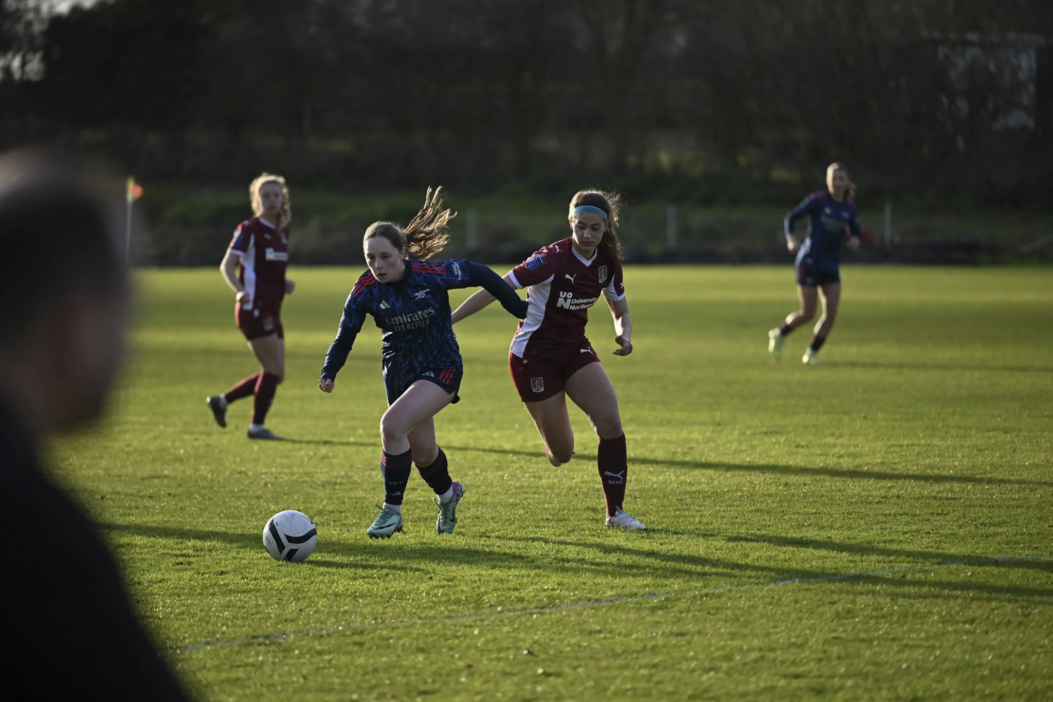 Women playing soccer on a grass field, with one woman in a blue jersey and another in a maroon jersey chasing the ball, while two other women are in the background.