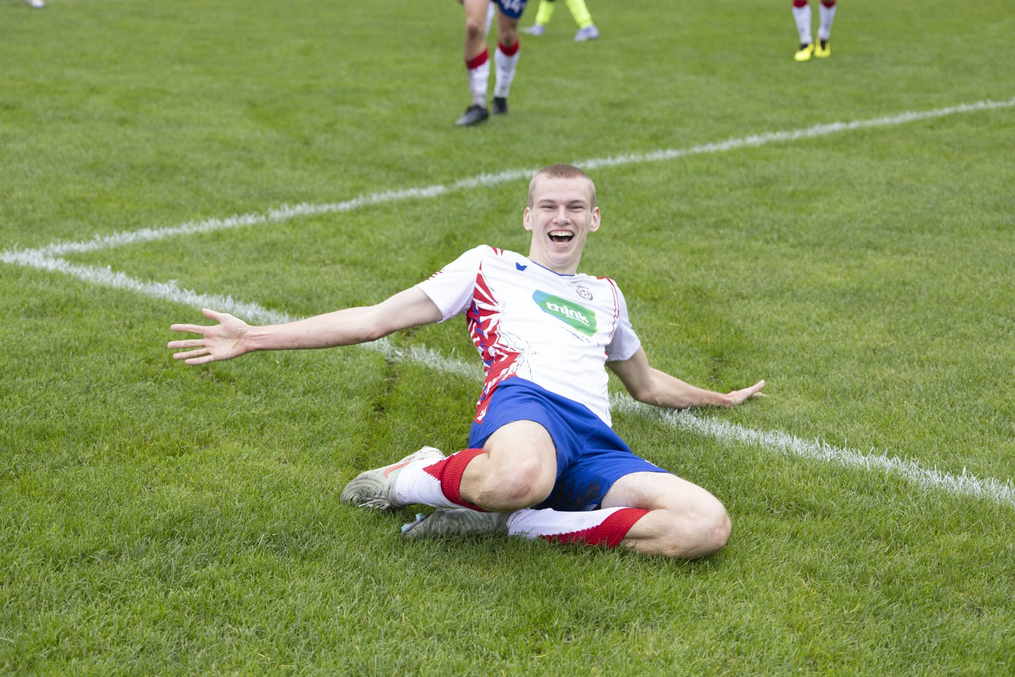A young soccer player celebrating on the field after scoring a goal, sitting on the grass with arms wide open and smiling.