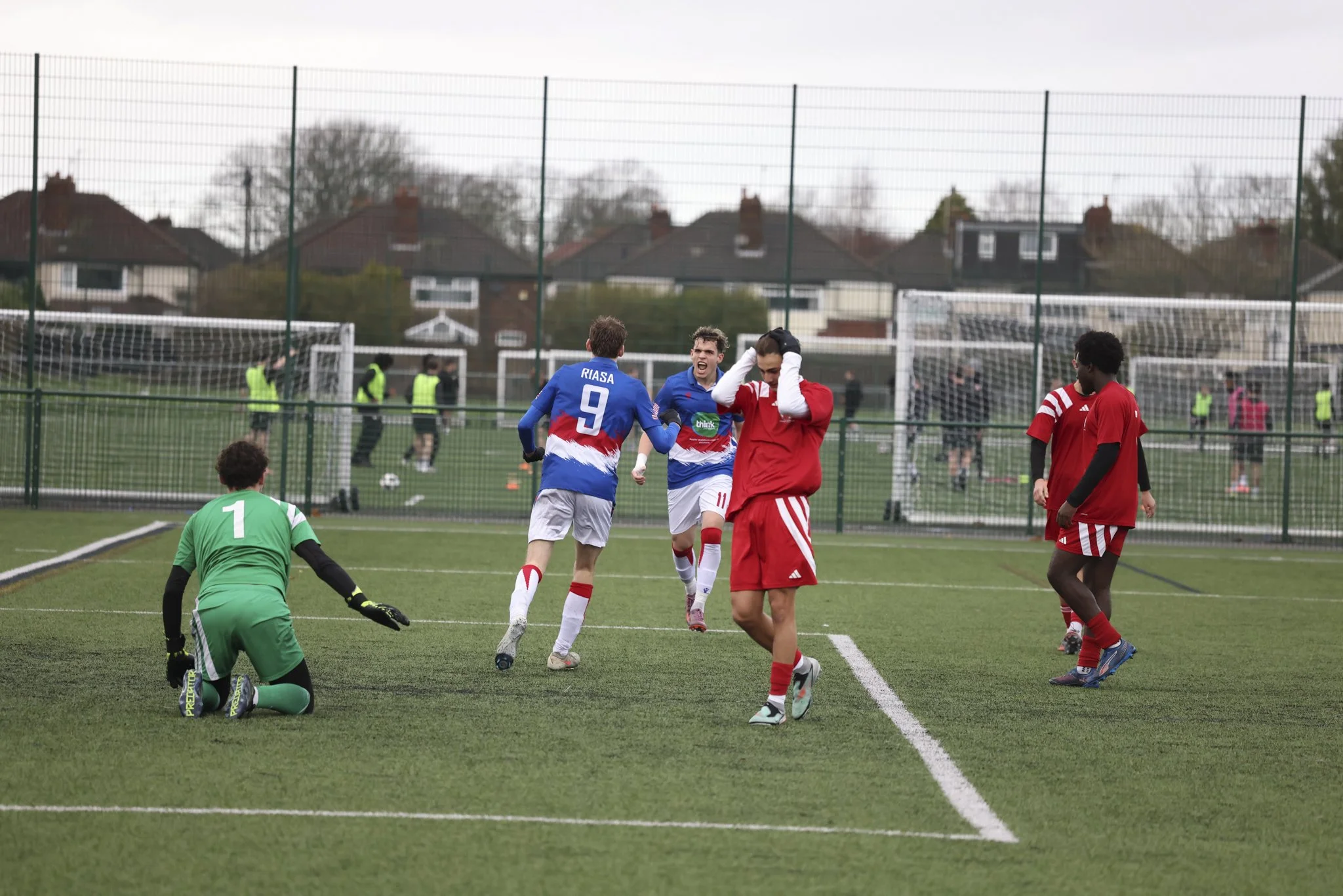 Soccer players on the field, some celebrating, others disappointed, after a goal; a goalkeeper in green kneels on the ground, players in red and blue jerseys are visible.