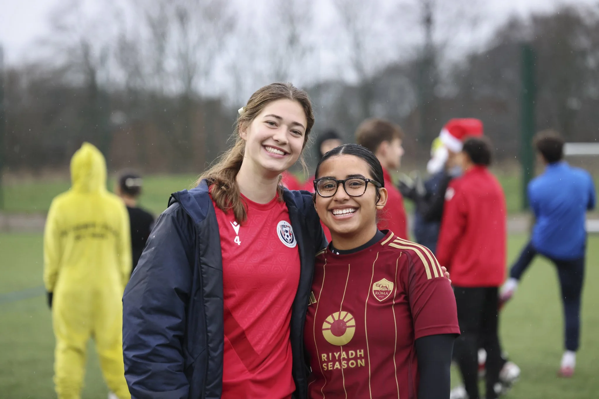 Two women smiling in soccer uniforms on a rainy outdoor soccer field, with other players in the background.