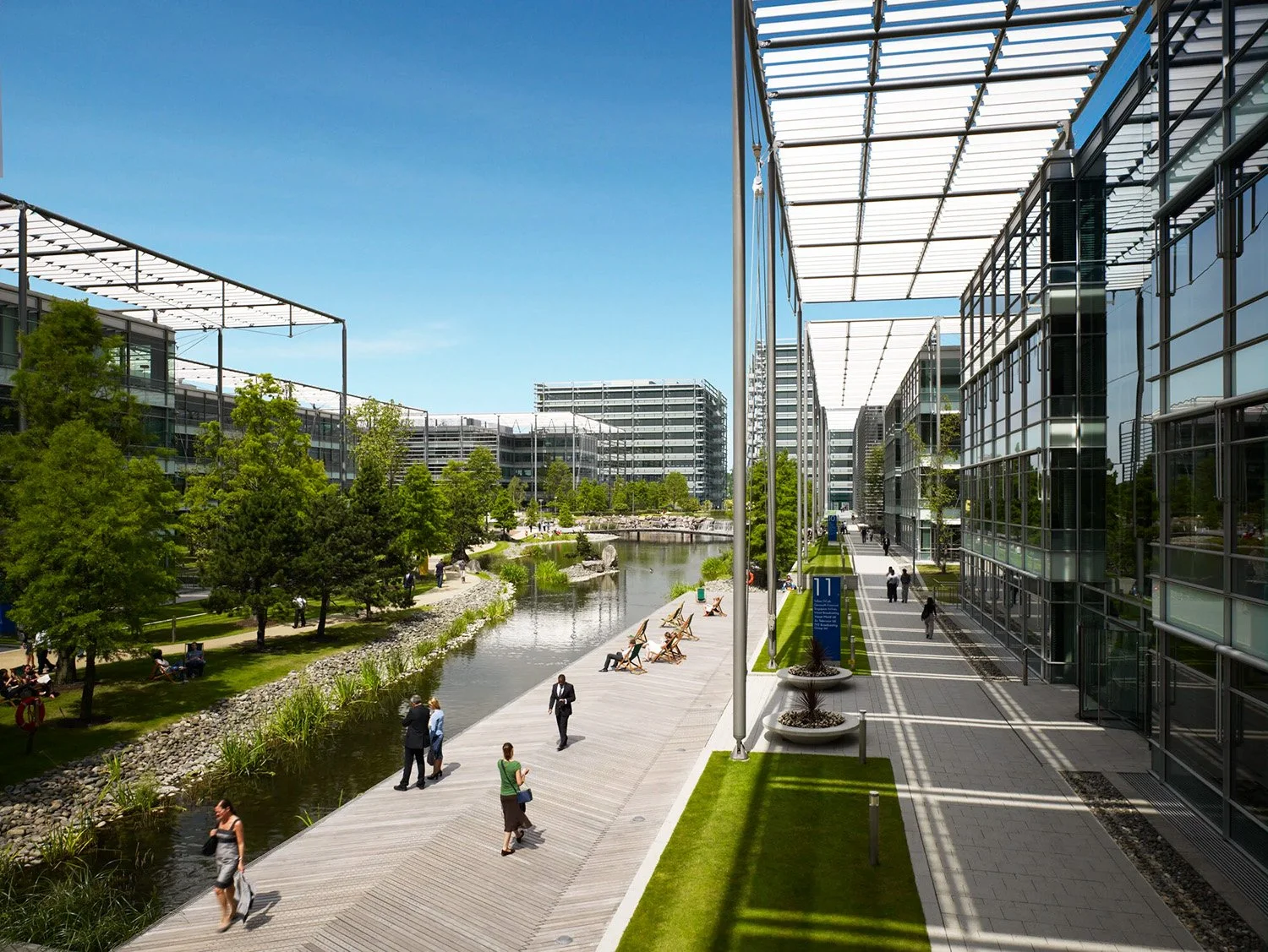 Modern office complex with glass buildings, a waterway, and green landscaping. People walk along the boardwalk, some sitting by the water, under a partially shaded structure.