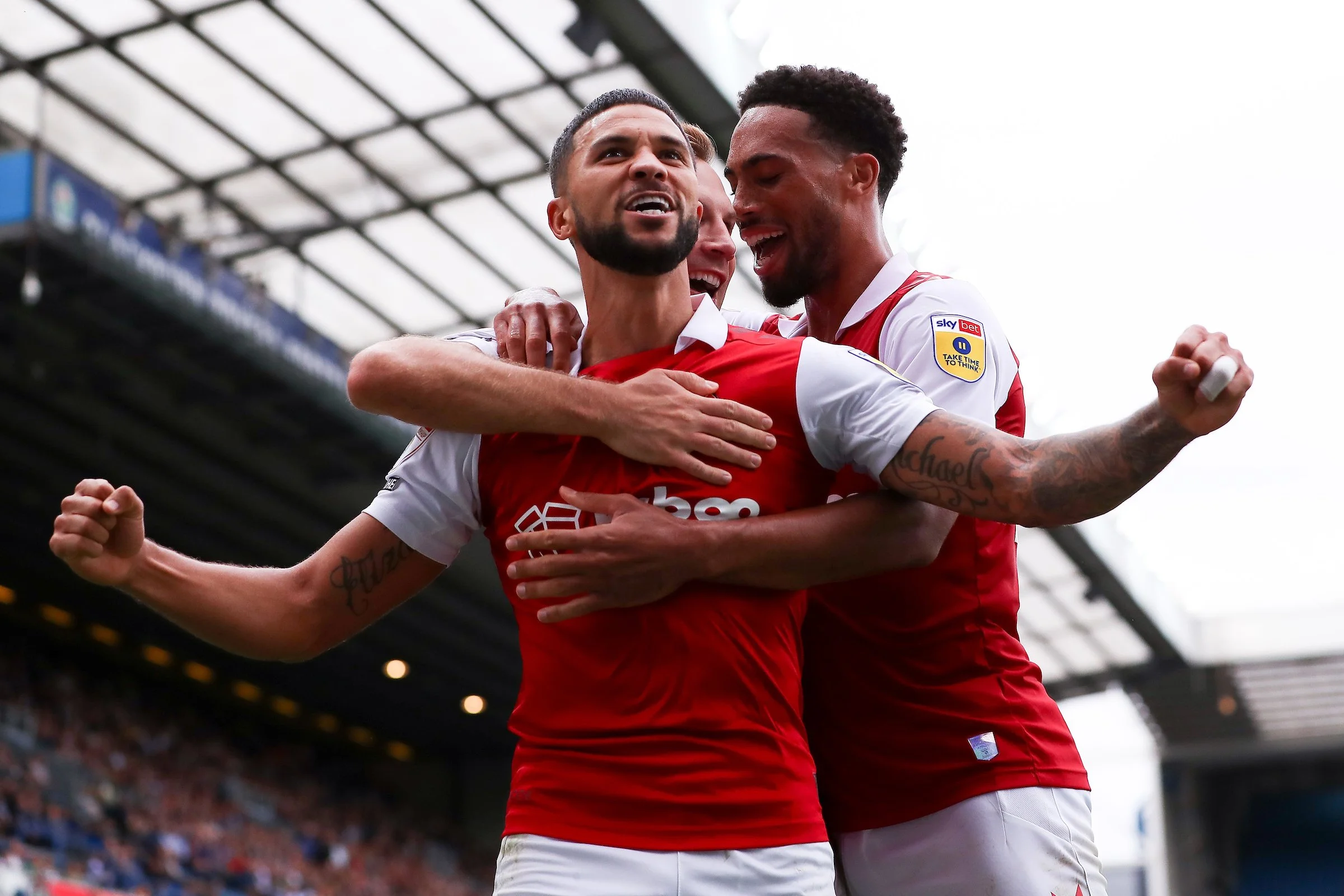 Three soccer players from Arsenal team celebrating on the field, wearing red and white uniforms, inside a stadium with a crowd in the background.