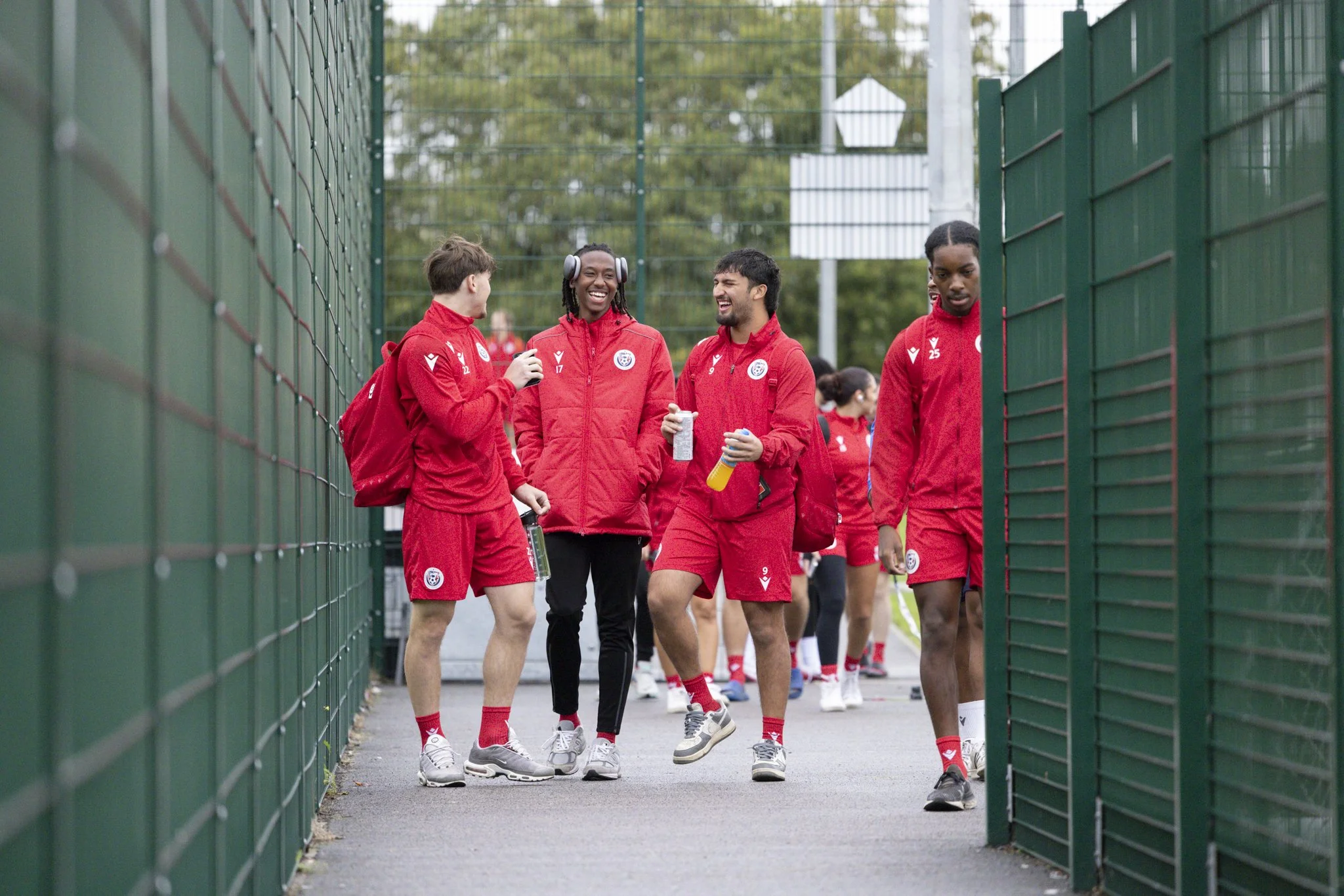 A group of female soccer players in red uniforms walking and talking on a path enclosed by green fences, some of them smiling, with a partially visible court or field in the background.