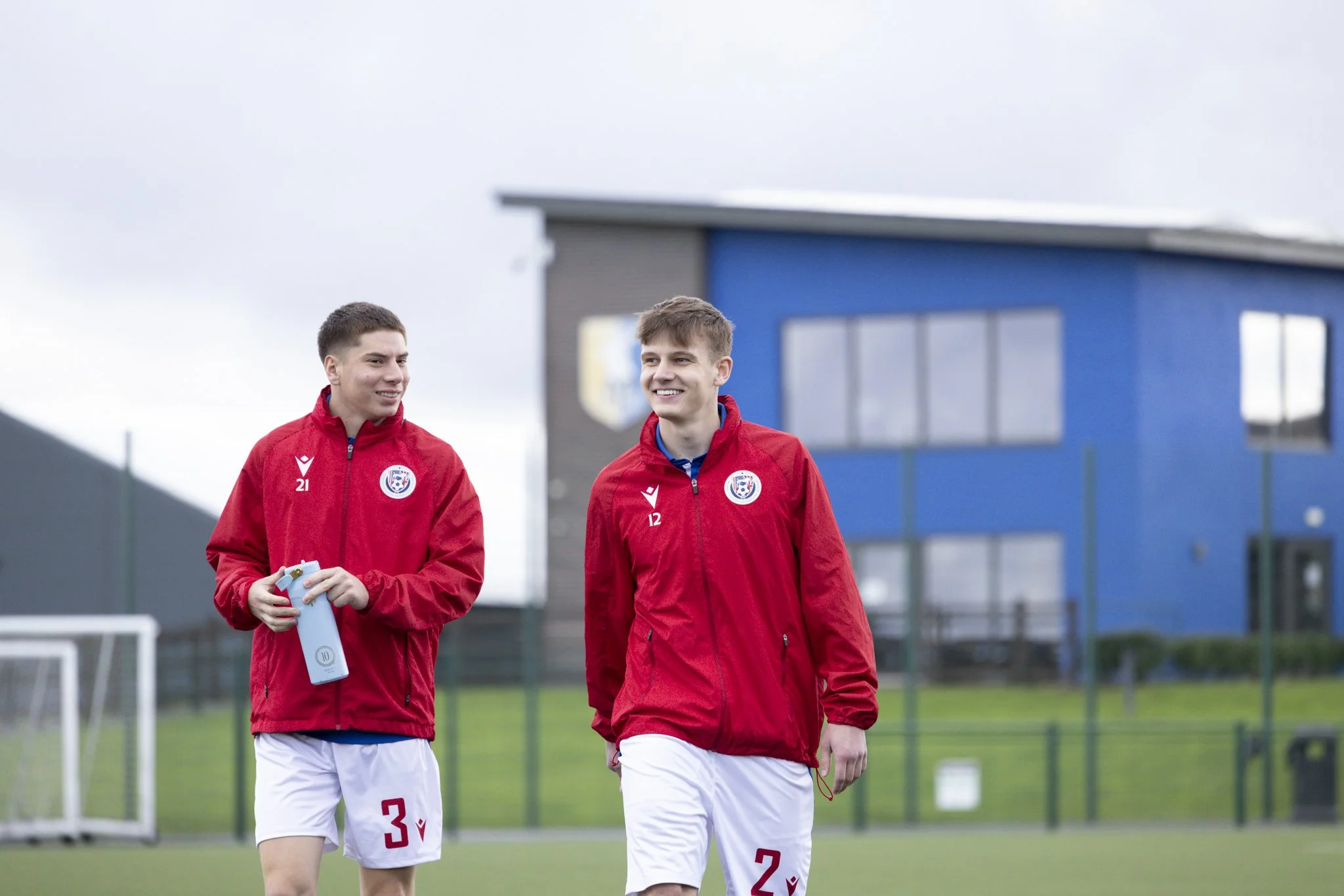 Two young male athletes wearing red sports jackets and white shorts, walking on a football field. One holds a water bottle, and both are smiling. The background features a blue building and a cloudy sky.