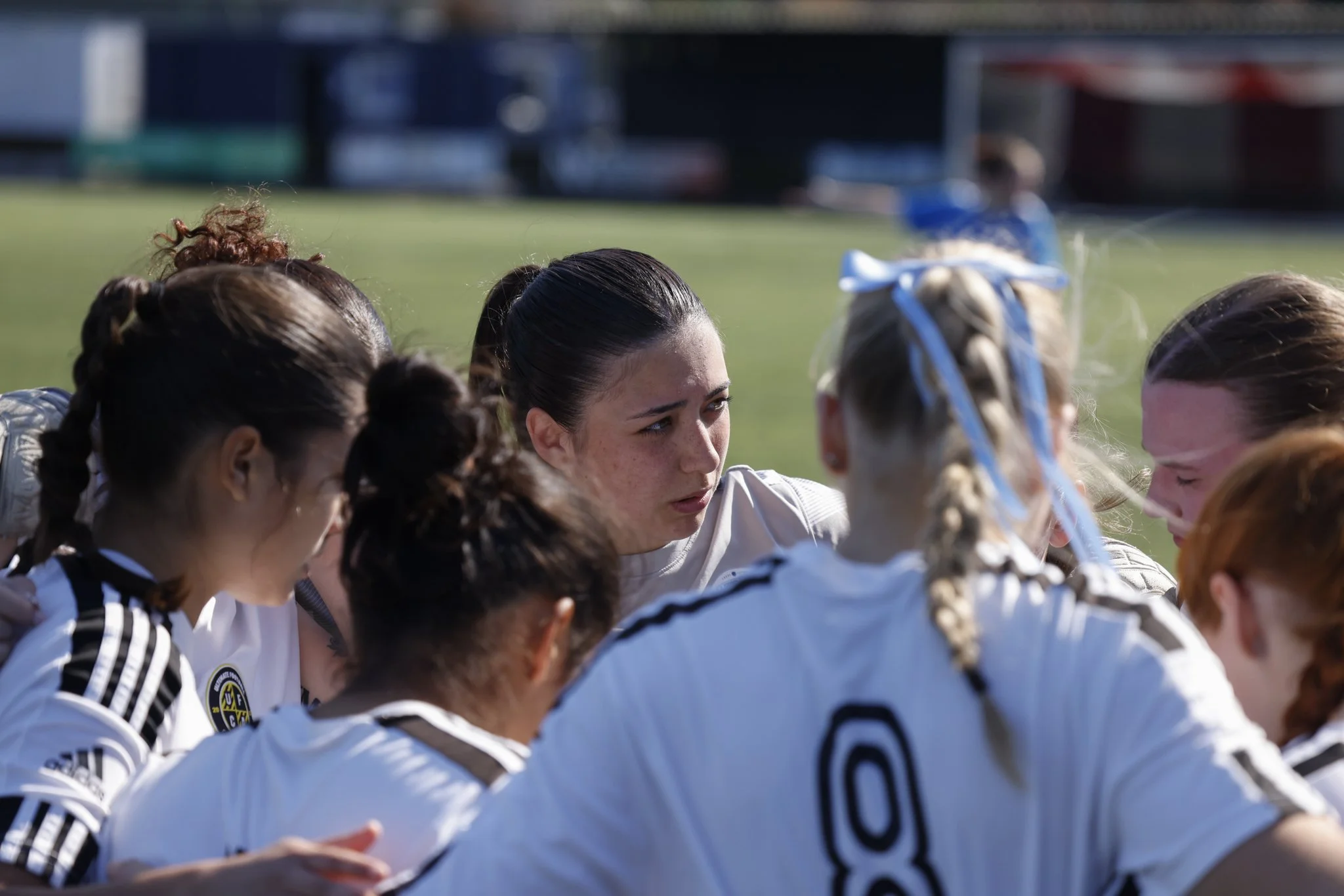 Female soccer players sit in a huddle on the field during a game, listening to the coach.