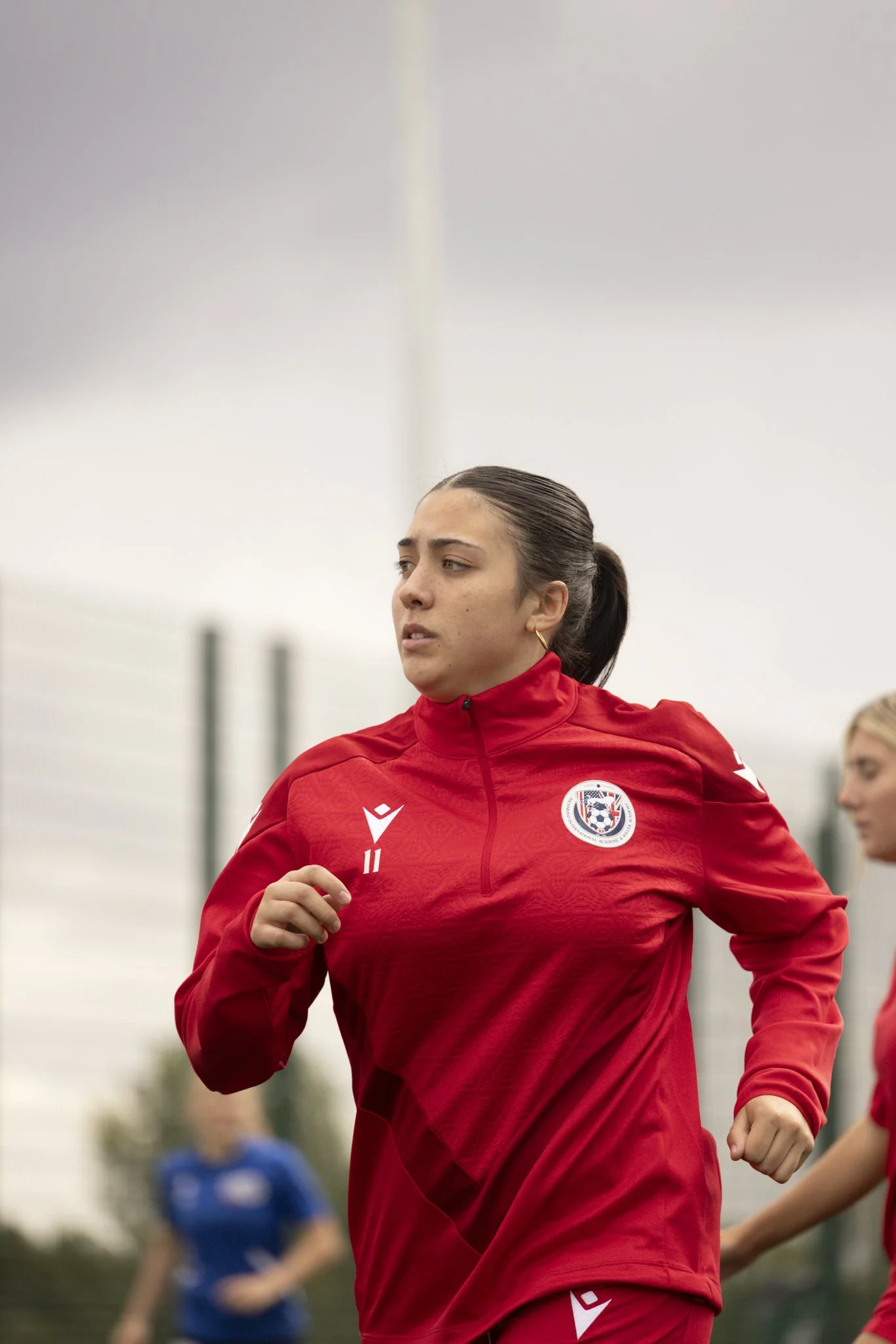A female soccer player wearing a red jacket with a team logo, jogging on a field.