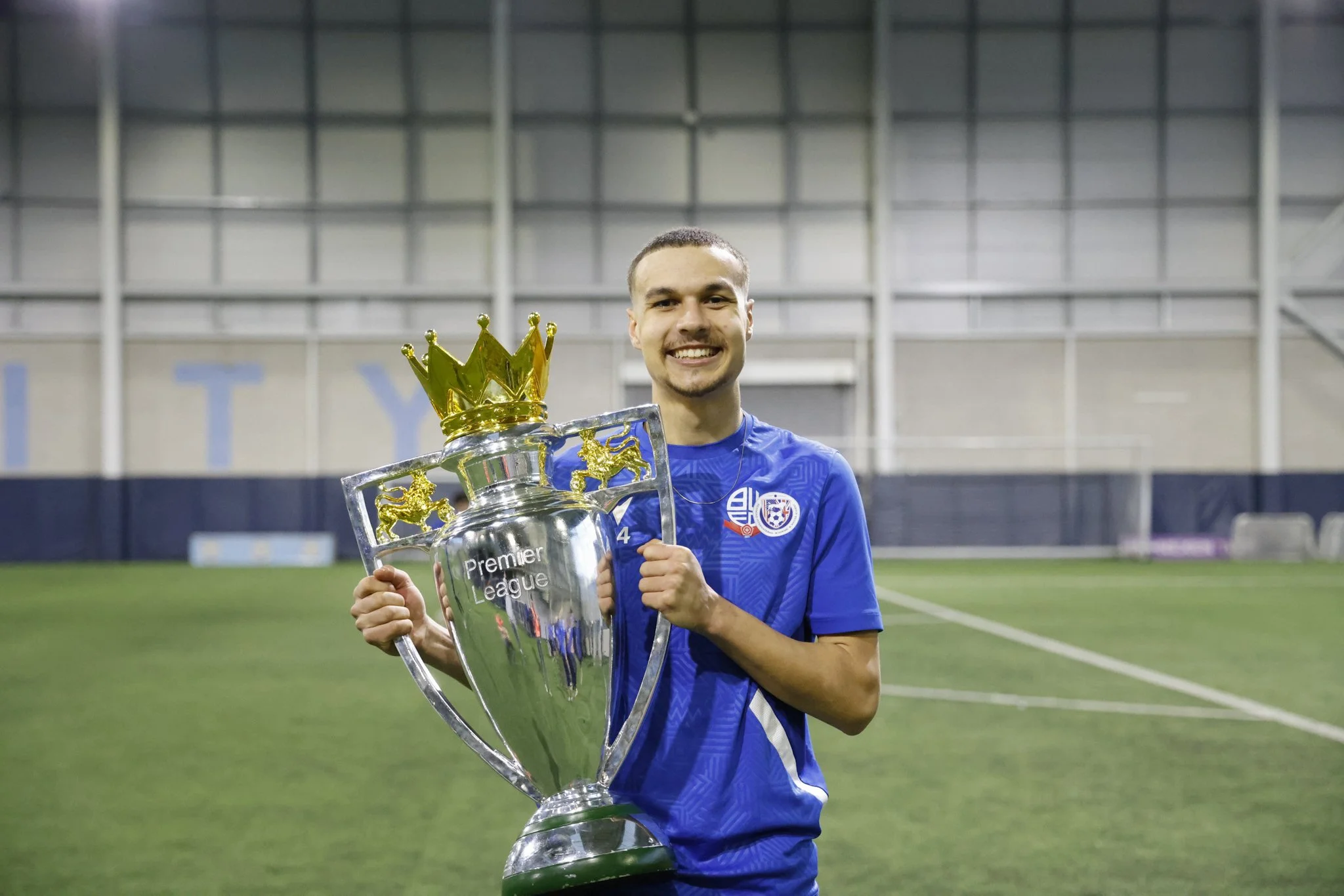 A young man in a blue soccer jersey holding a large trophy inside a sports facility
