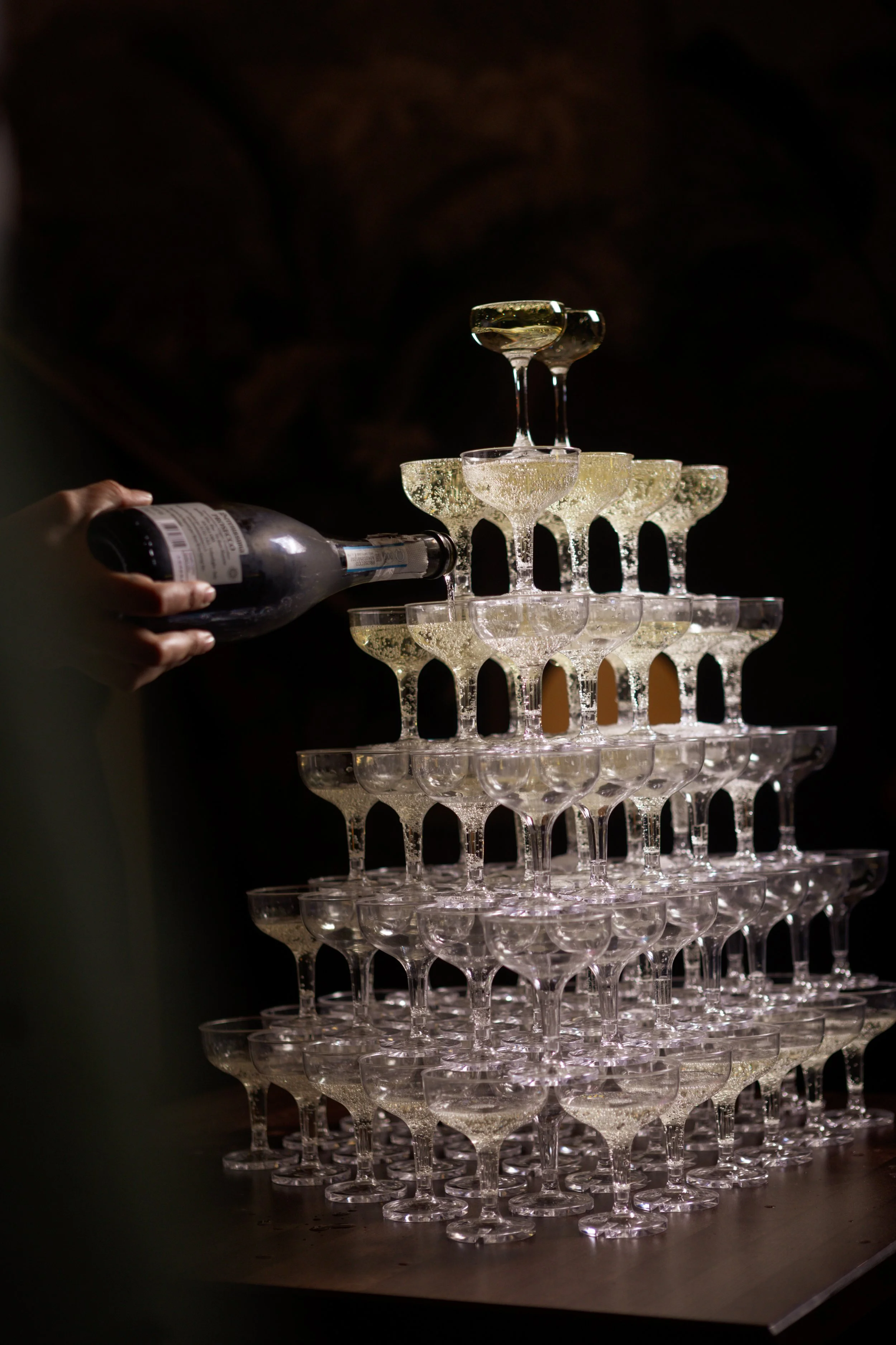 A person pours champagne into a stacked pyramid of champagne glasses.