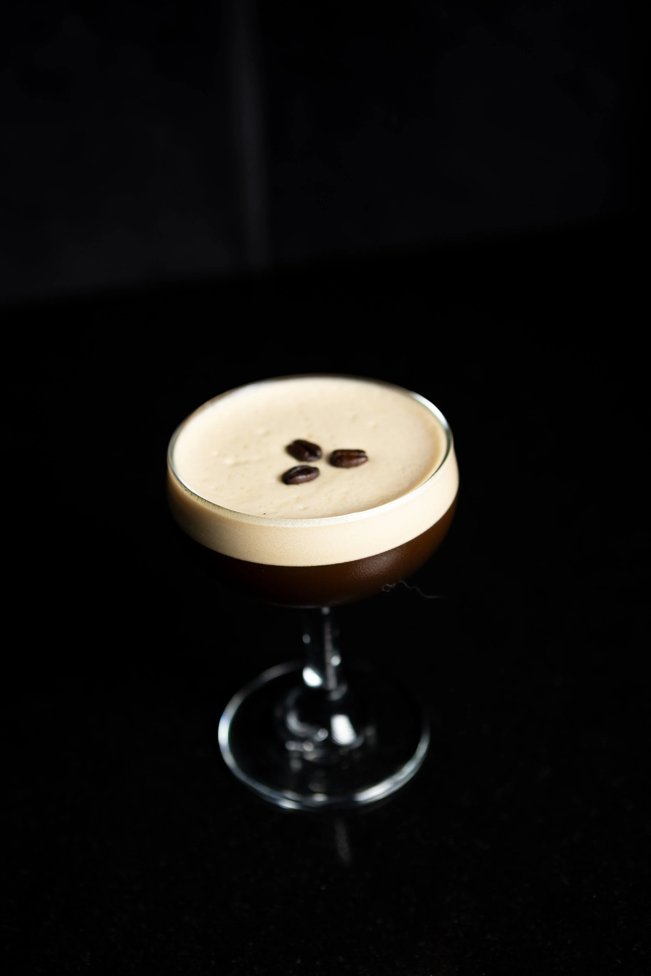 A cocktail drink in a coupe glass with a dark base, topped with a creamy foam and garnished with three coffee beans, set against a black background.