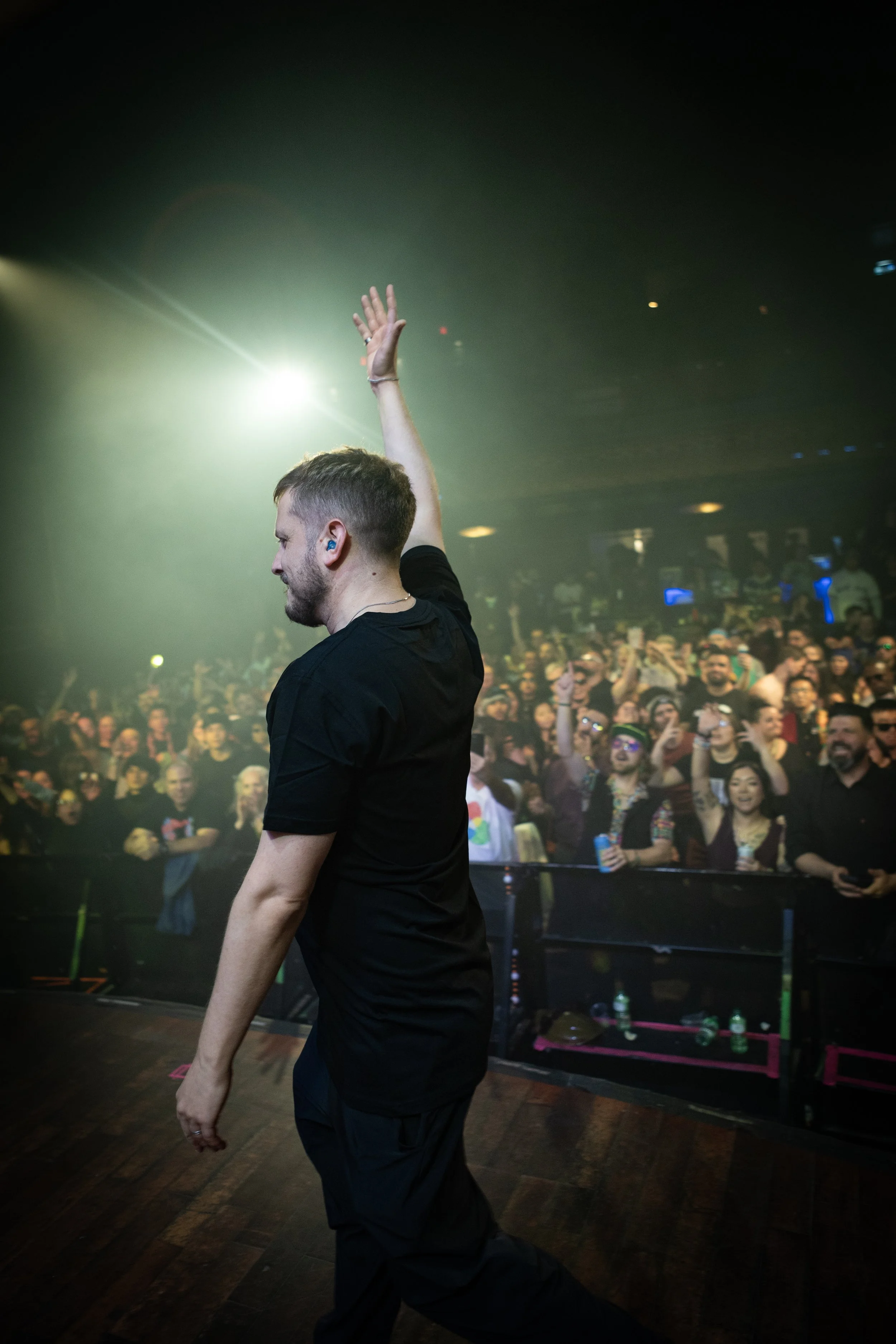A DJ with short hair and a beard raising his hand on stage, performing at a concert with a large, diverse audience cheering in the background.
