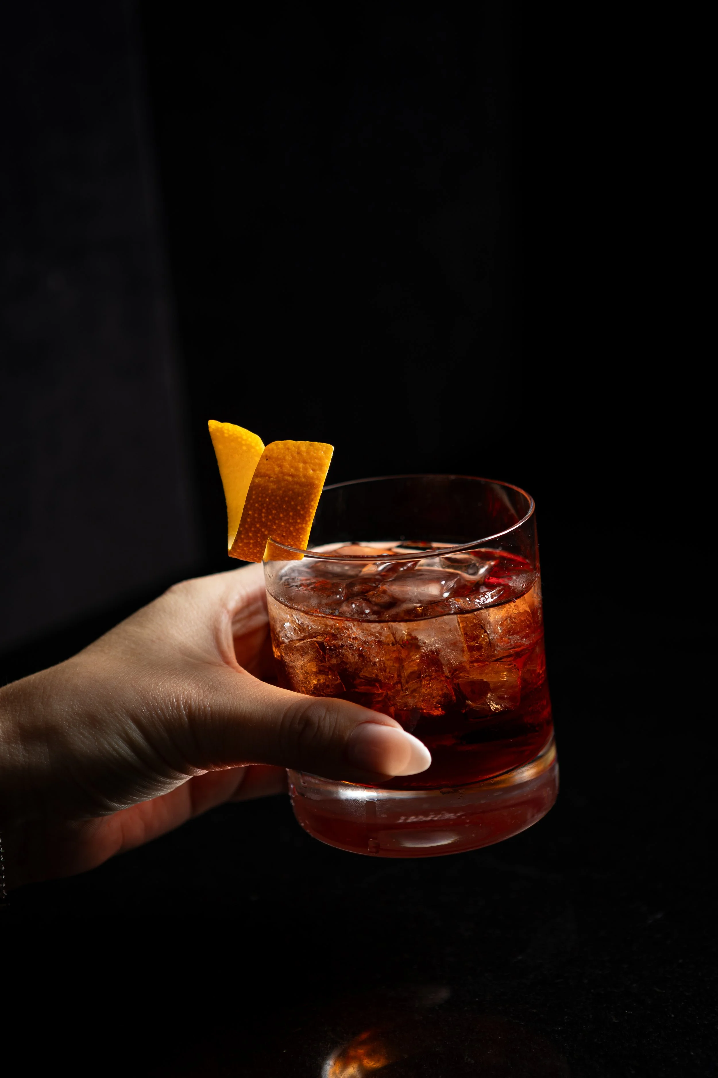 Close-up of a hand holding a glass of red cocktail with ice, garnished with a twist of lemon or orange peel, against a black background.